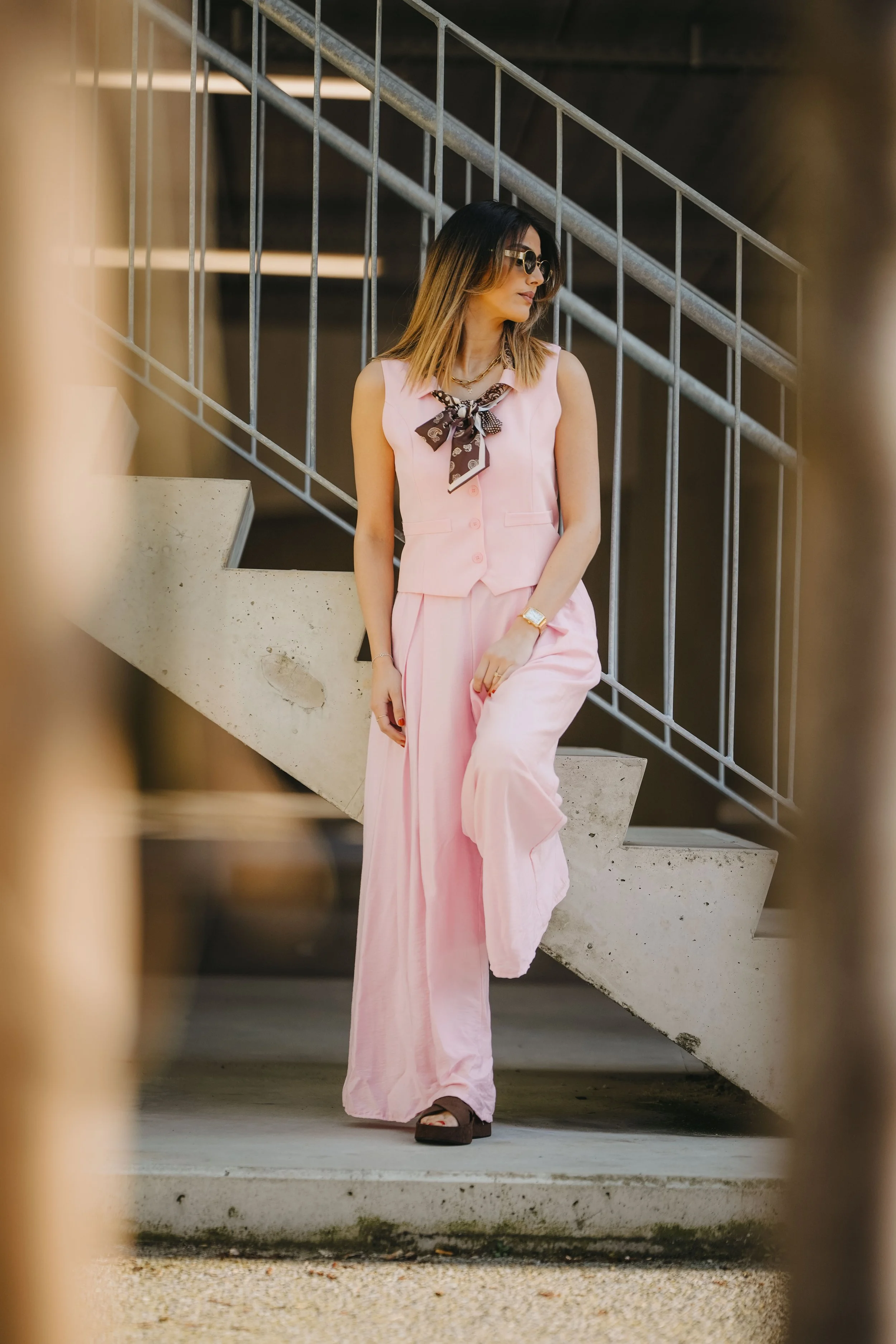 Femme portant un ensemble rose pastel composé d'une veste sans manches et d'un pantalon, avec un foulard à motifs autour du cou, posée sur un escalier en béton, vue de profile.