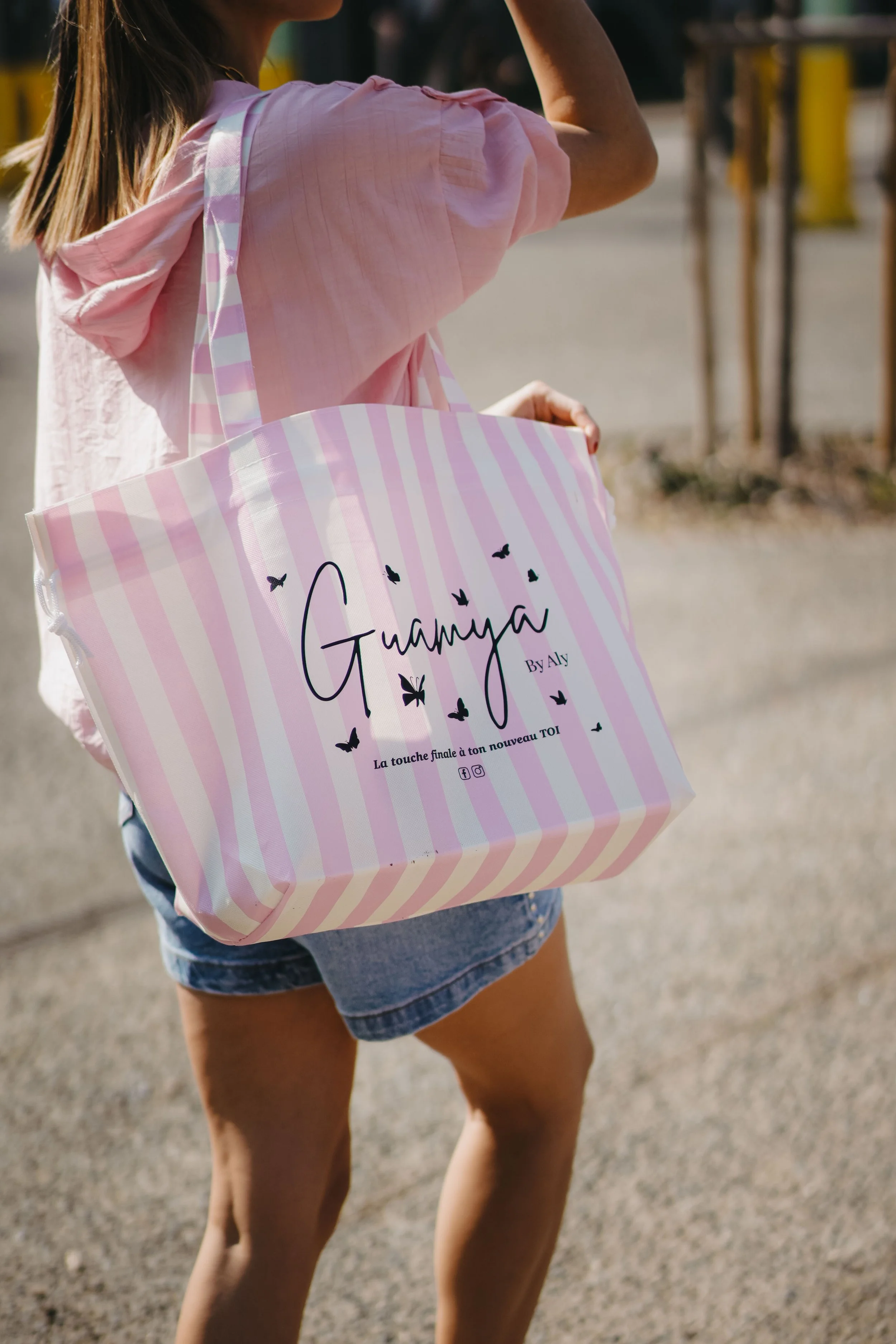 Une femme porte un sac de plage rose à rayures avec le texte "Gauguin by Aly" et des papillons noirs, en dehors, dans une zone en plein air.