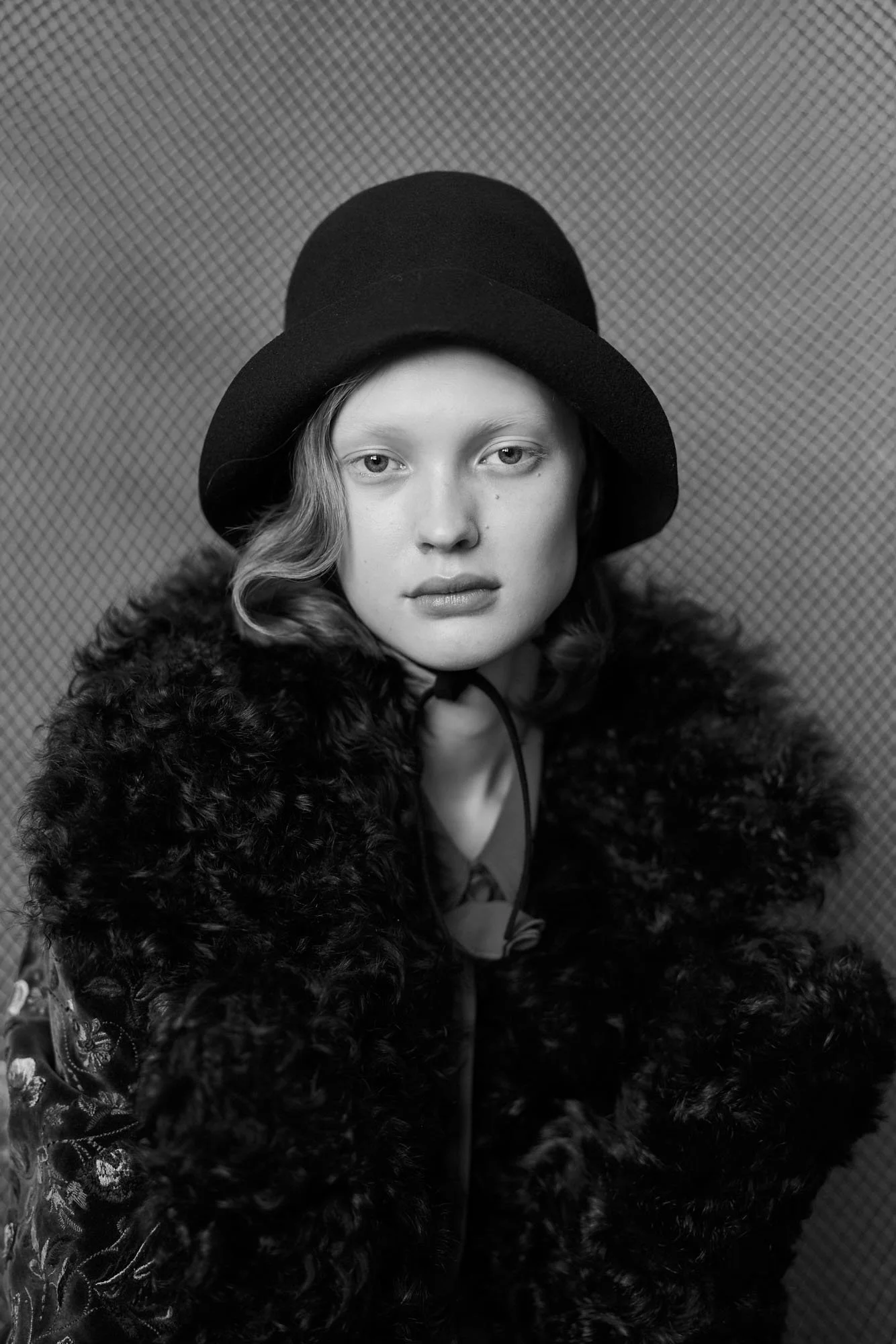 Schwarz-Weiß-Foto eines jungen Mädchens mit dele Akzent, das einen dunklen Hut und einen Pelzmantel trägt, vor einer texturierten Wand.
