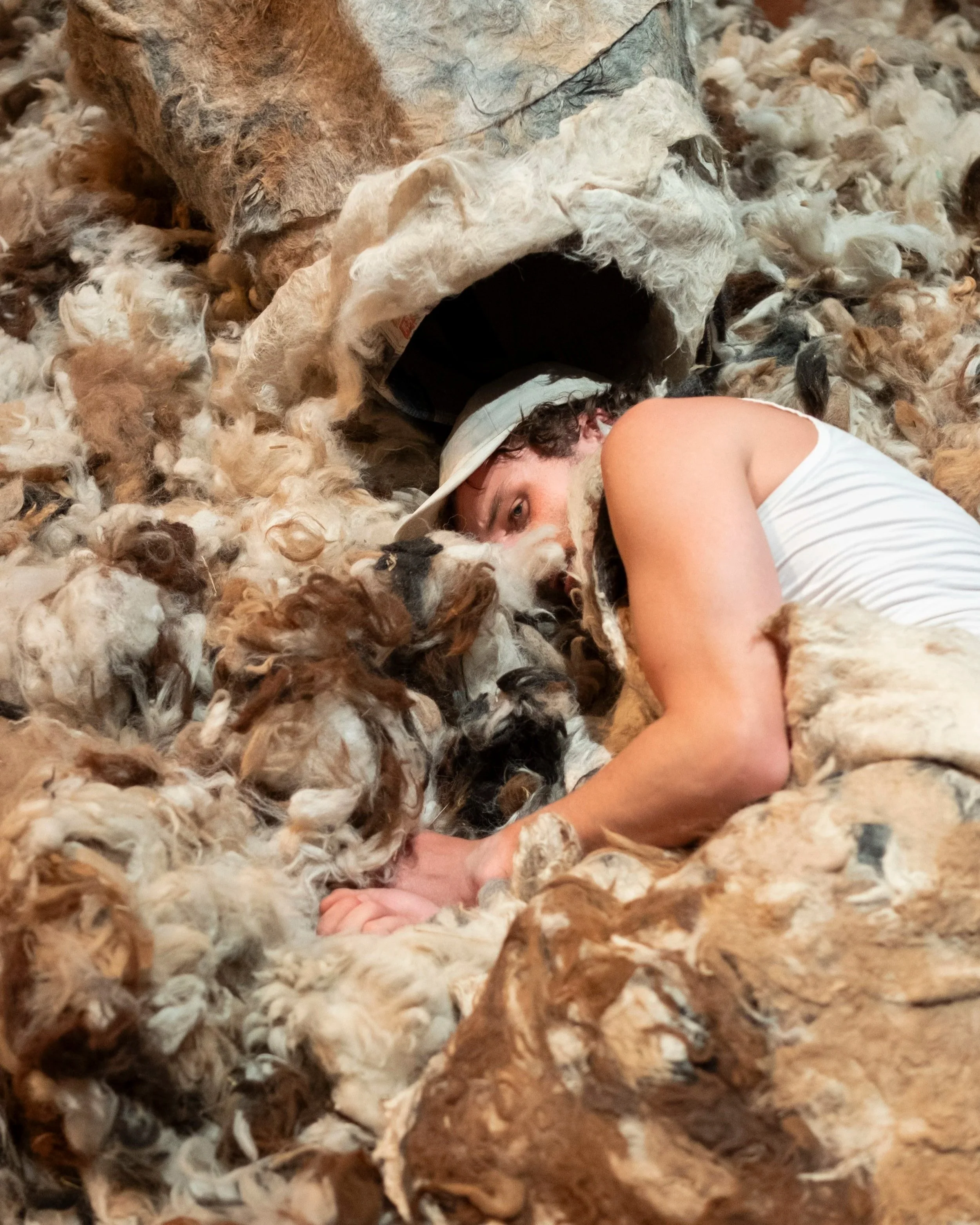 noah janssen ligt onder een pet tussen een grote hoeveelheid alpacawol. boven hem ligt een door valentijn schmitz en nico pul gemaakt vilten masker. 