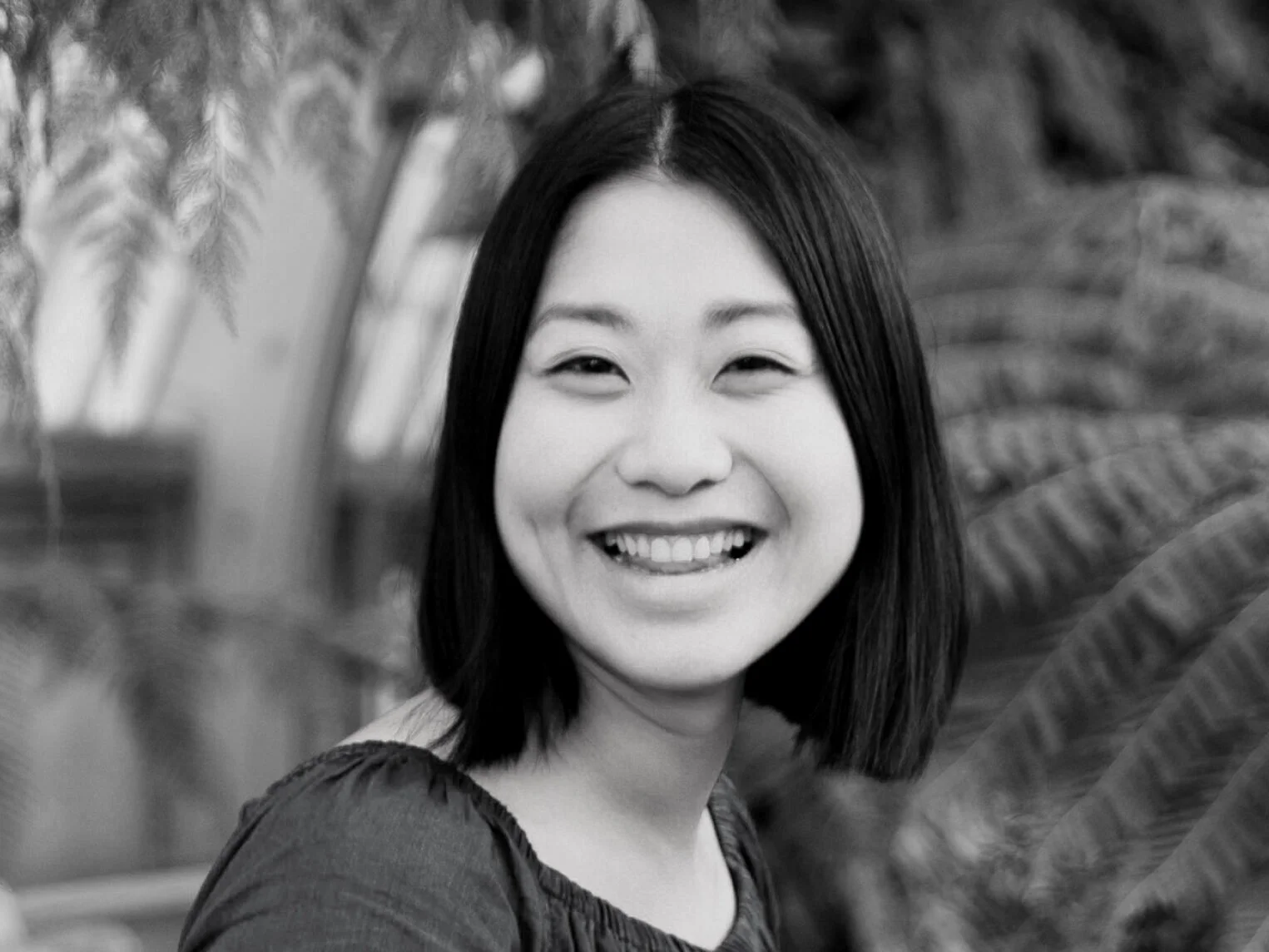 A smiling woman with shoulder-length black hair in a garden or greenhouse with plants and large leaves around her.