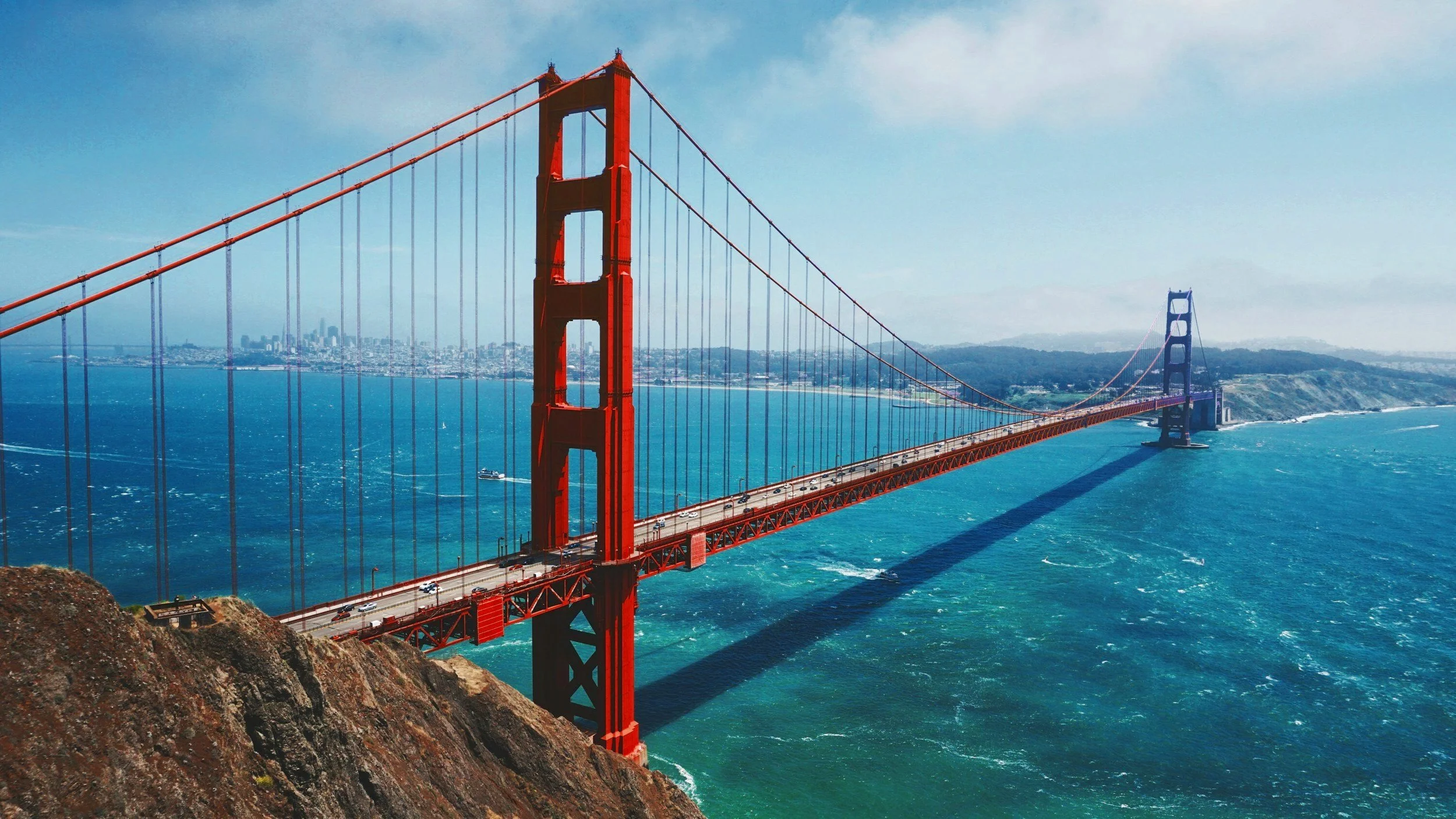 Golden Gate Bridge spanning across blue ocean waters with city skyline in the background, clear sky overhead.