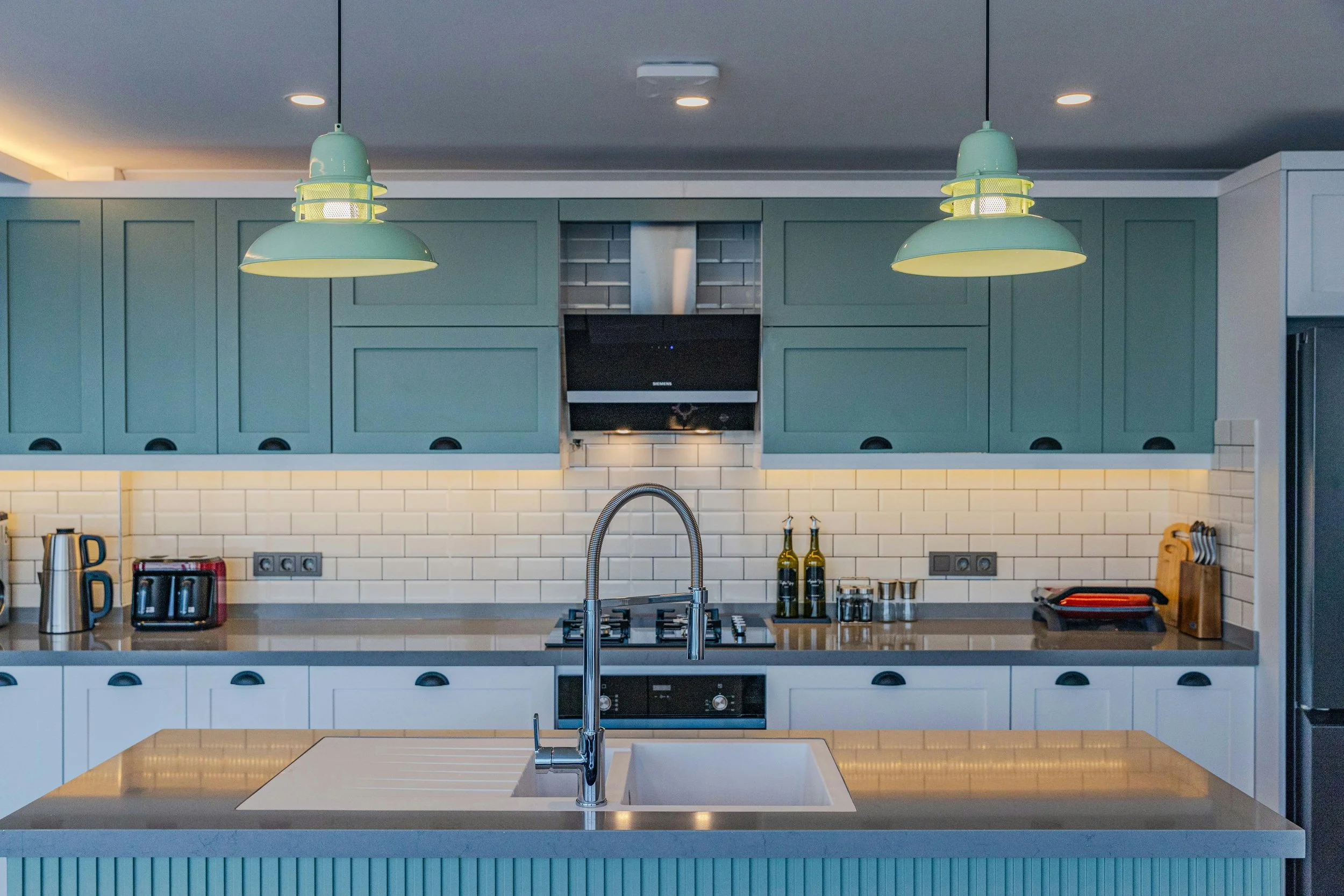 Modern kitchen with mint green cabinets, white subway tiles, two pendant lights, stainless steel appliances, and a central island with a sink.