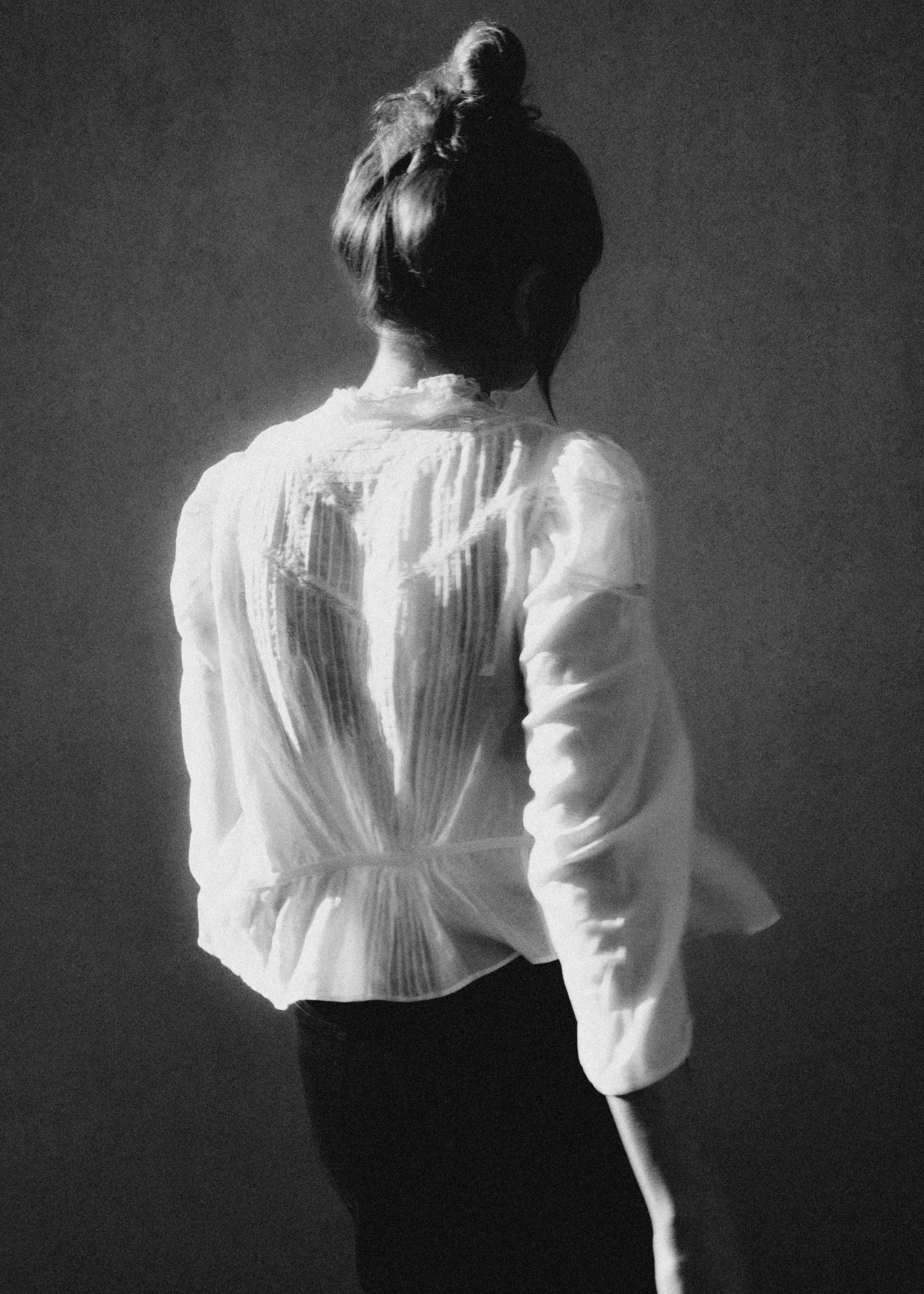 A person with their back turned, wearing a light-colored blouse, with hair tied up in a bun, holding a flower against a dark background.