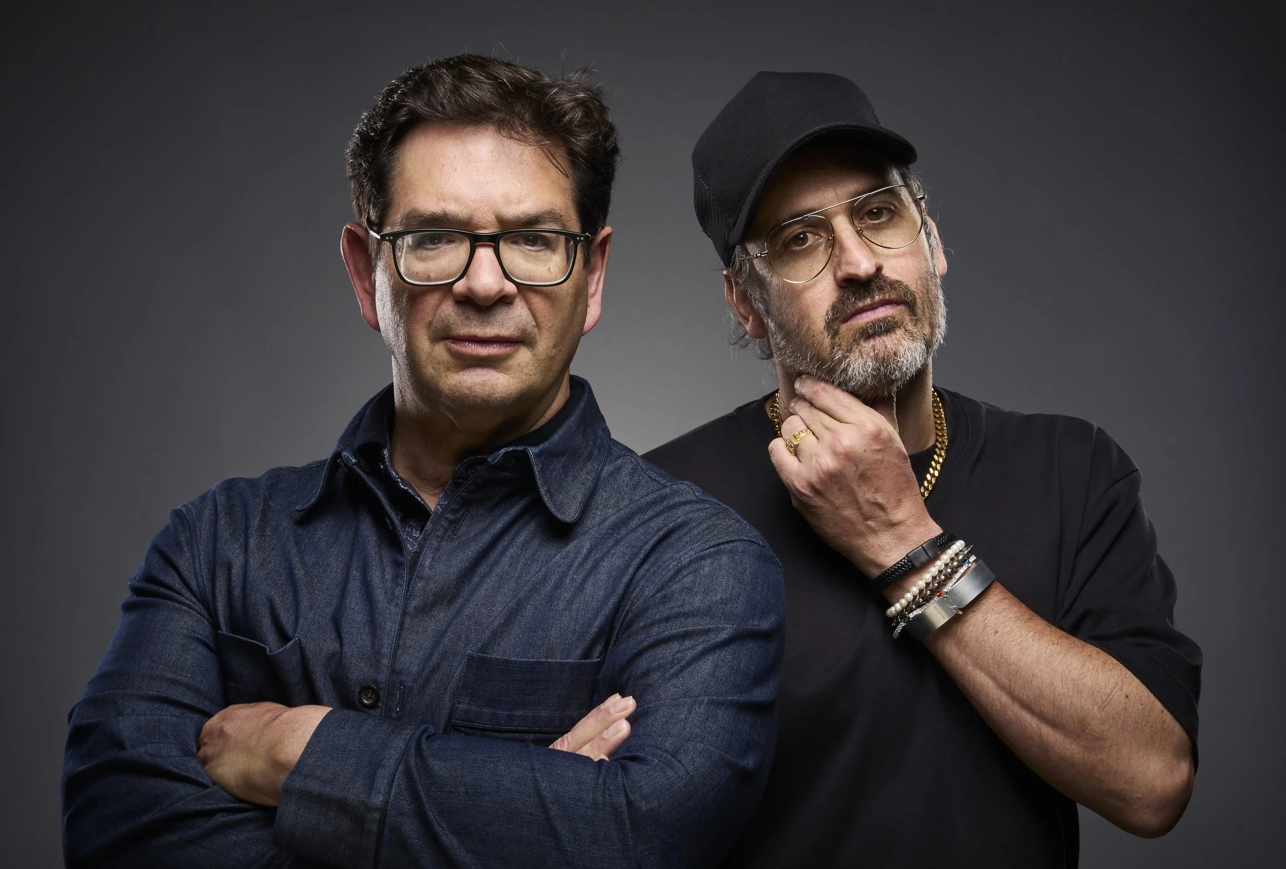 Two men standing against a dark background, one wearing glasses, a denim jacket, and crossing his arms, the other wearing glasses, a black cap, and touching his chin with his hand, dressed in black.