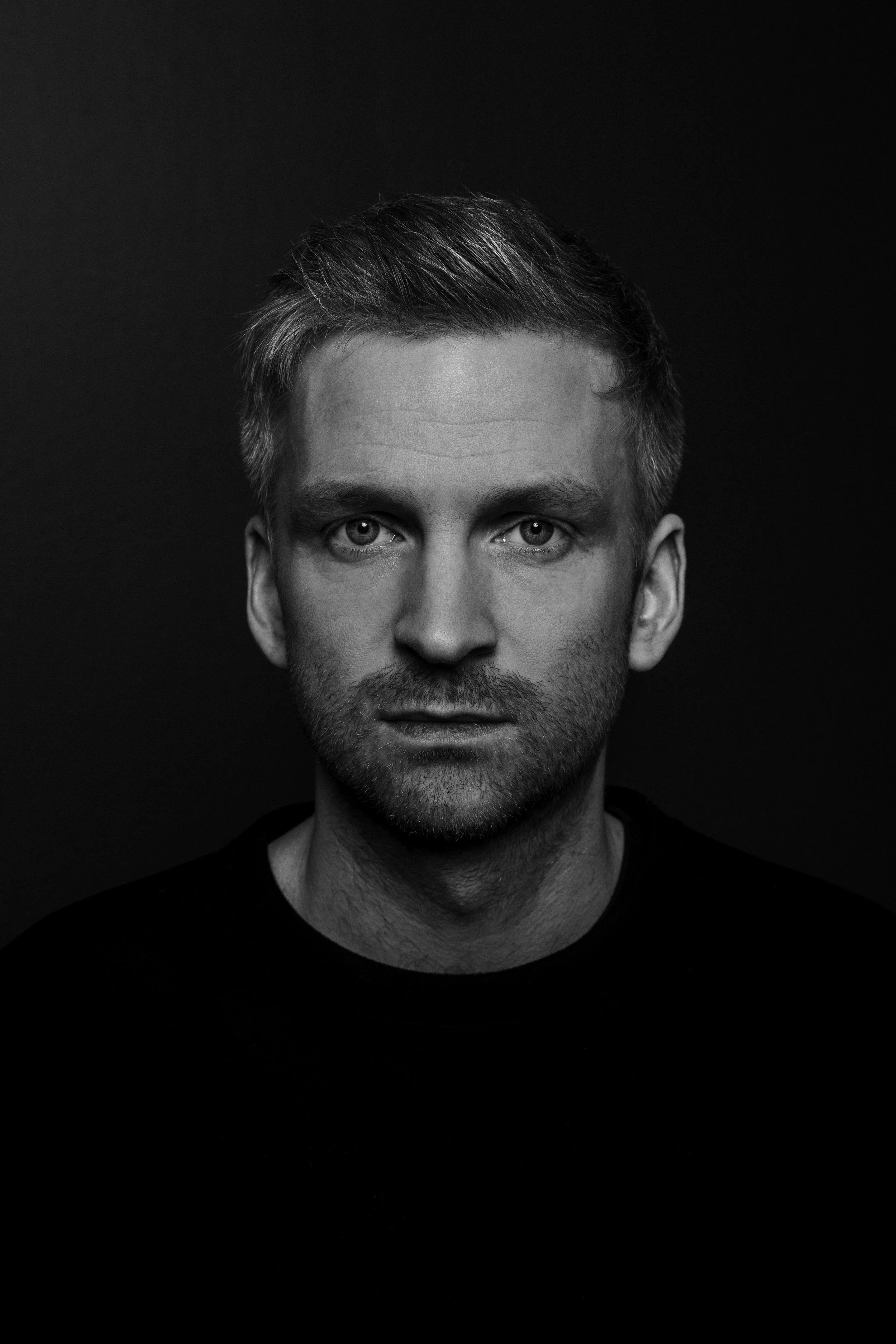 Black and white portrait of a young man with light-colored hair and a slight beard, looking directly at the camera, dark background.