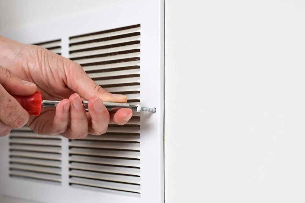A person using a screwdriver to screw a screw into a white vent cover.