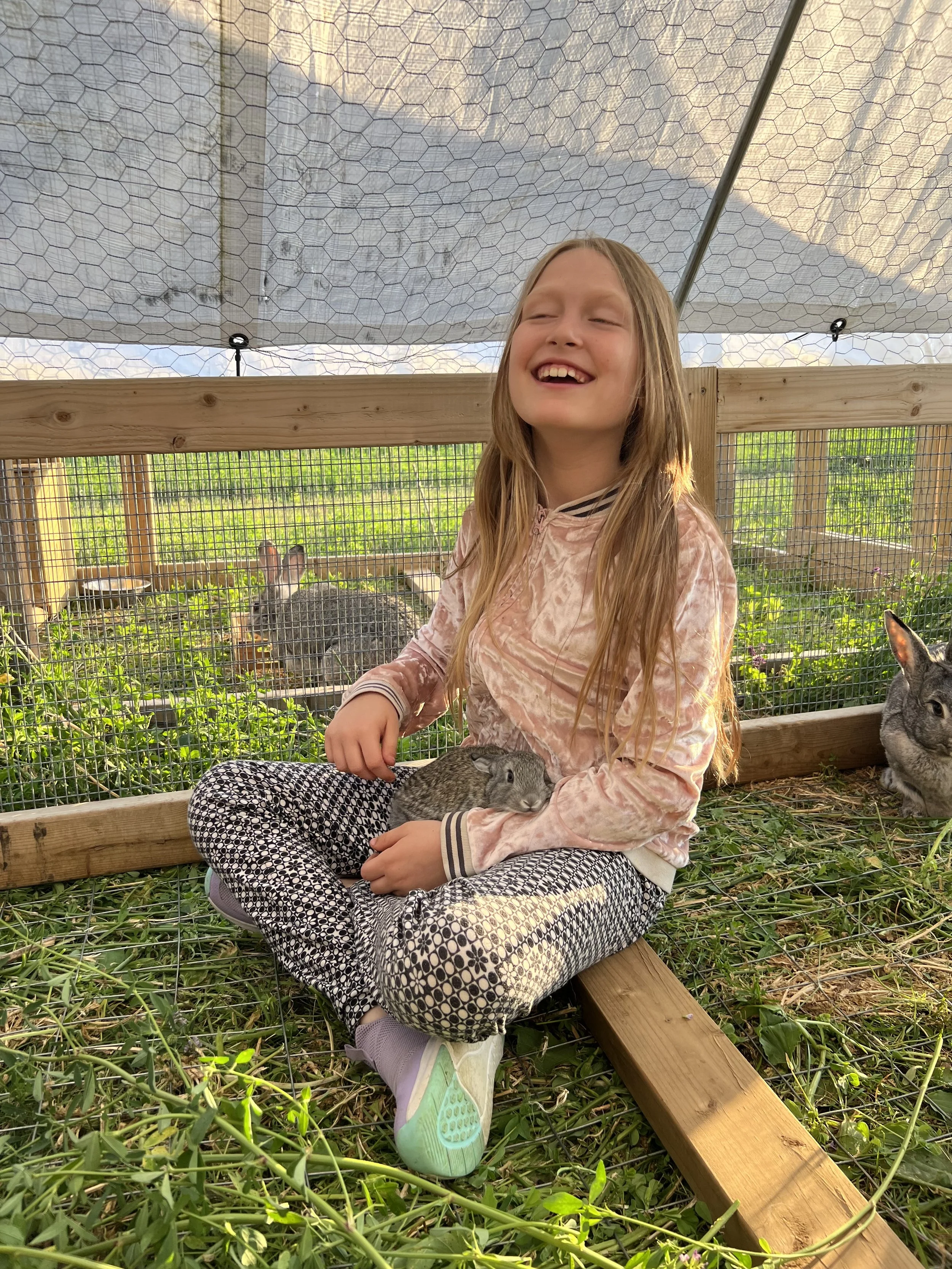 A happy girl with long blonde hair, wearing a pink velvet jacket, patterned leggings, and sneakers, sitting cross-legged in a bunny enclosure, holding a small rabbit.