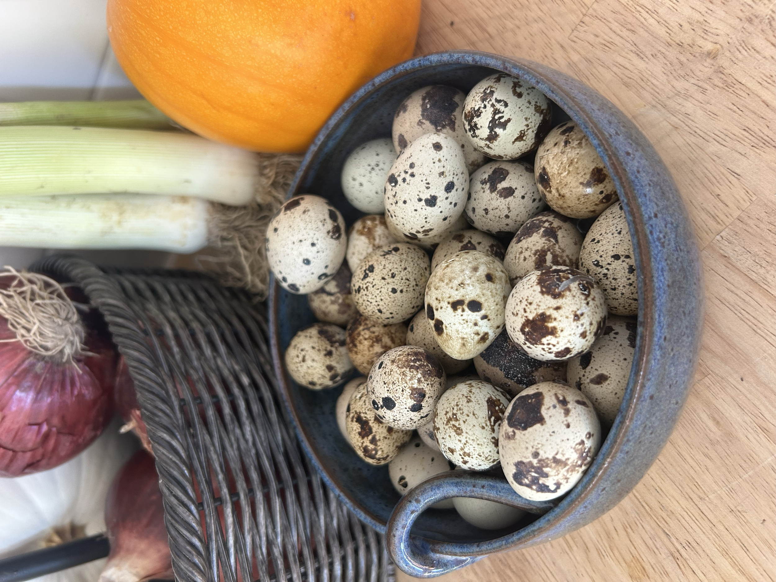 A group of quail eggs in a ceramic bowl, with a pumpkin, a leek, and onions nearby on a wooden surface.