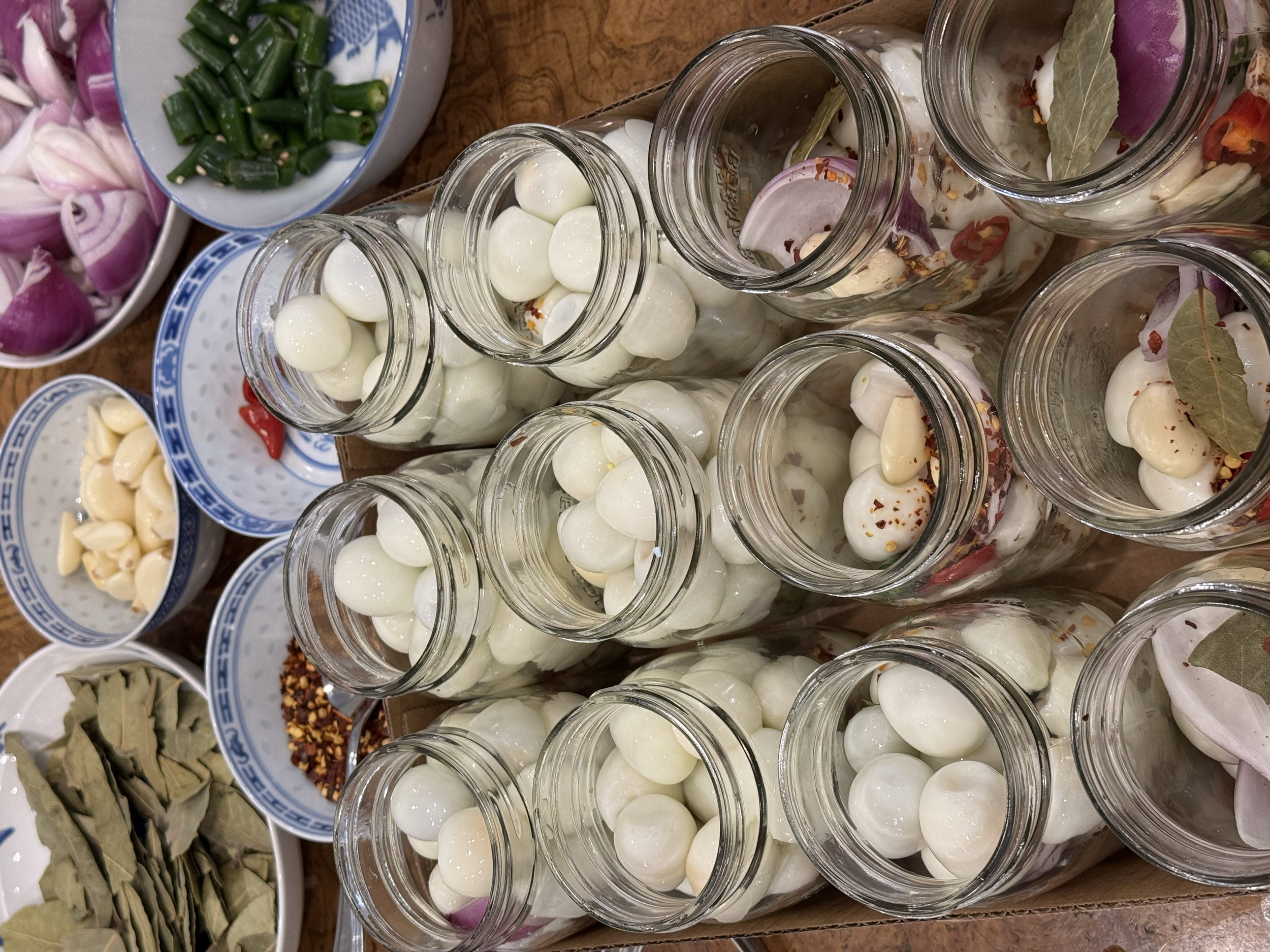 Multiple jars filled with white garlic cloves, surrounded by small bowls containing chopped green onions, garlic, chili flakes, and bay leaves.
