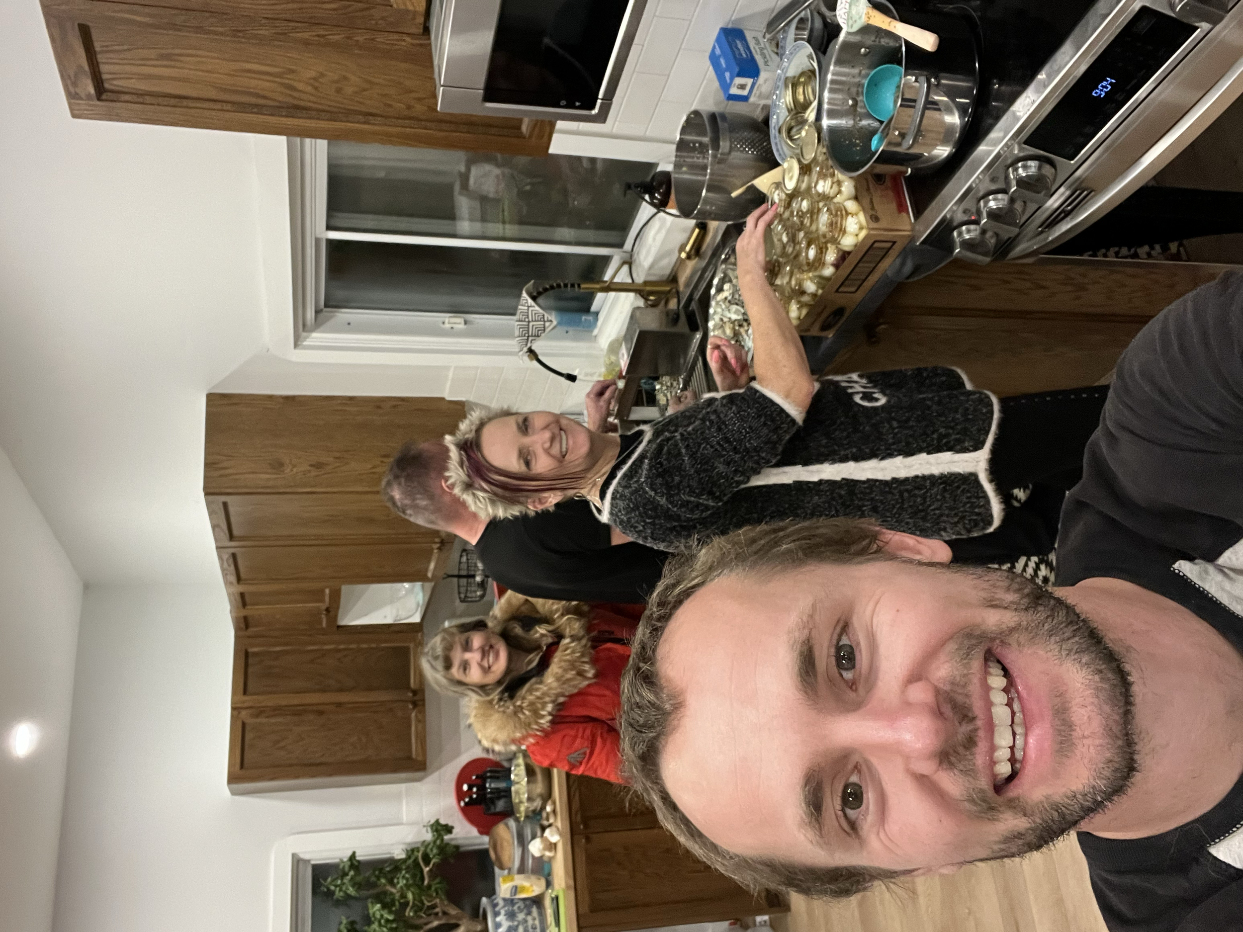 A group of three people in a kitchen preparing food, with one person taking a selfie. The kitchen has wooden cabinets, a window, and various cooking utensils and ingredients on the stove and countertops.