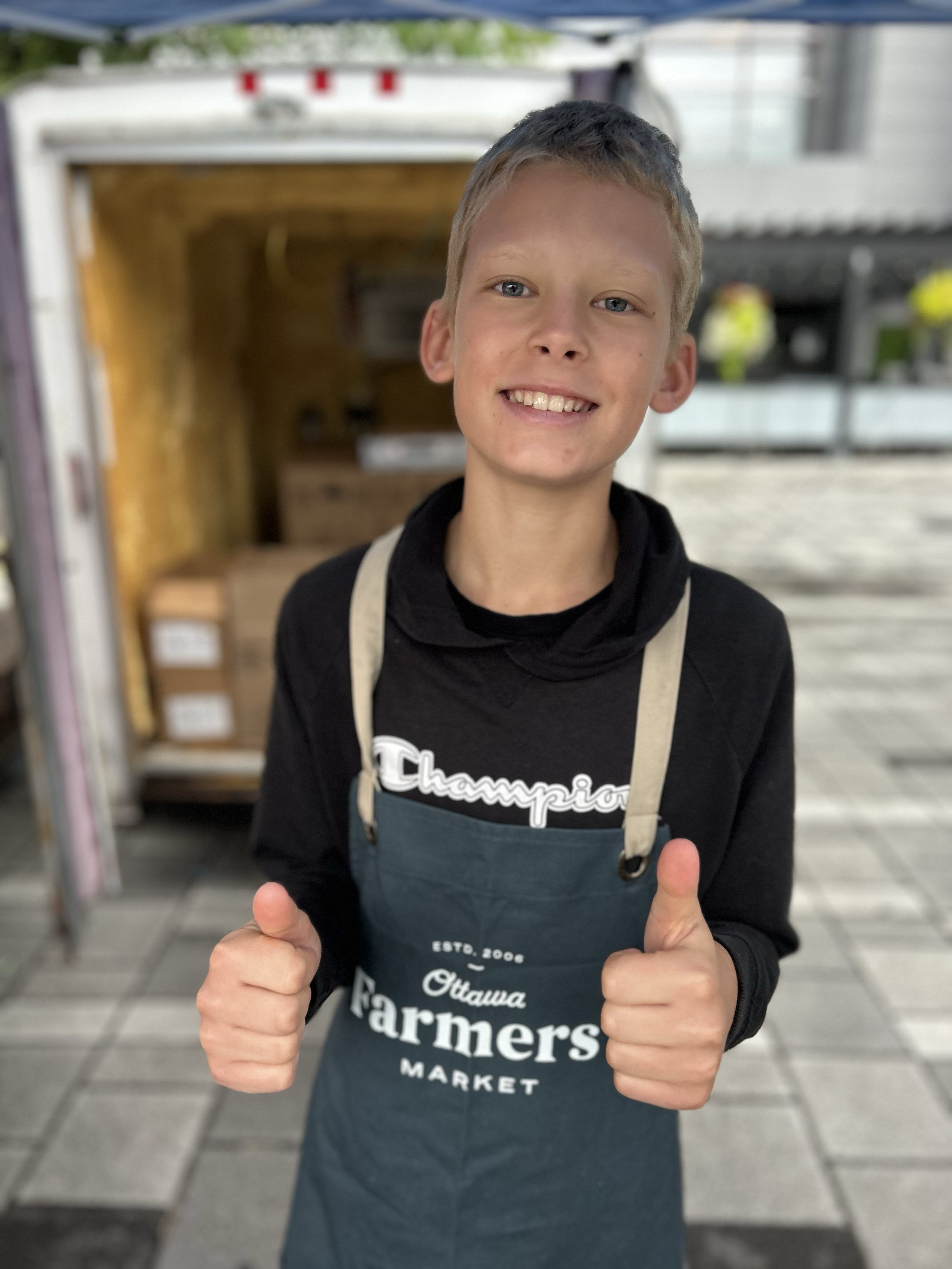 A young boy smiling and giving a thumbs-up, wearing an apron at the Ottawa Farmers Market.