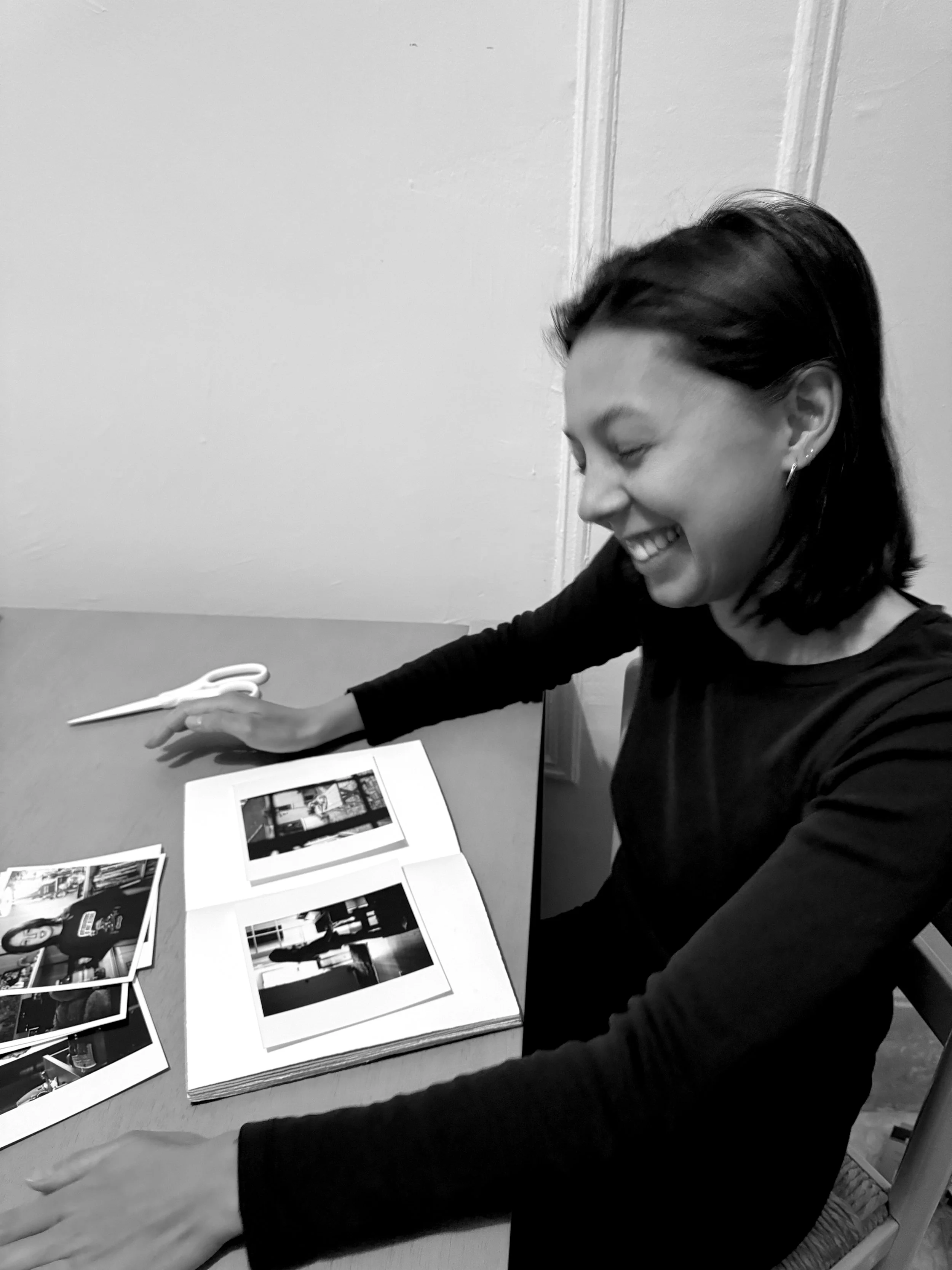 A woman sitting at a table, smiling and looking down at a photo album with photographs on a white page. There are additional photographs scattered on the table, and a pair of scissors lies nearby.