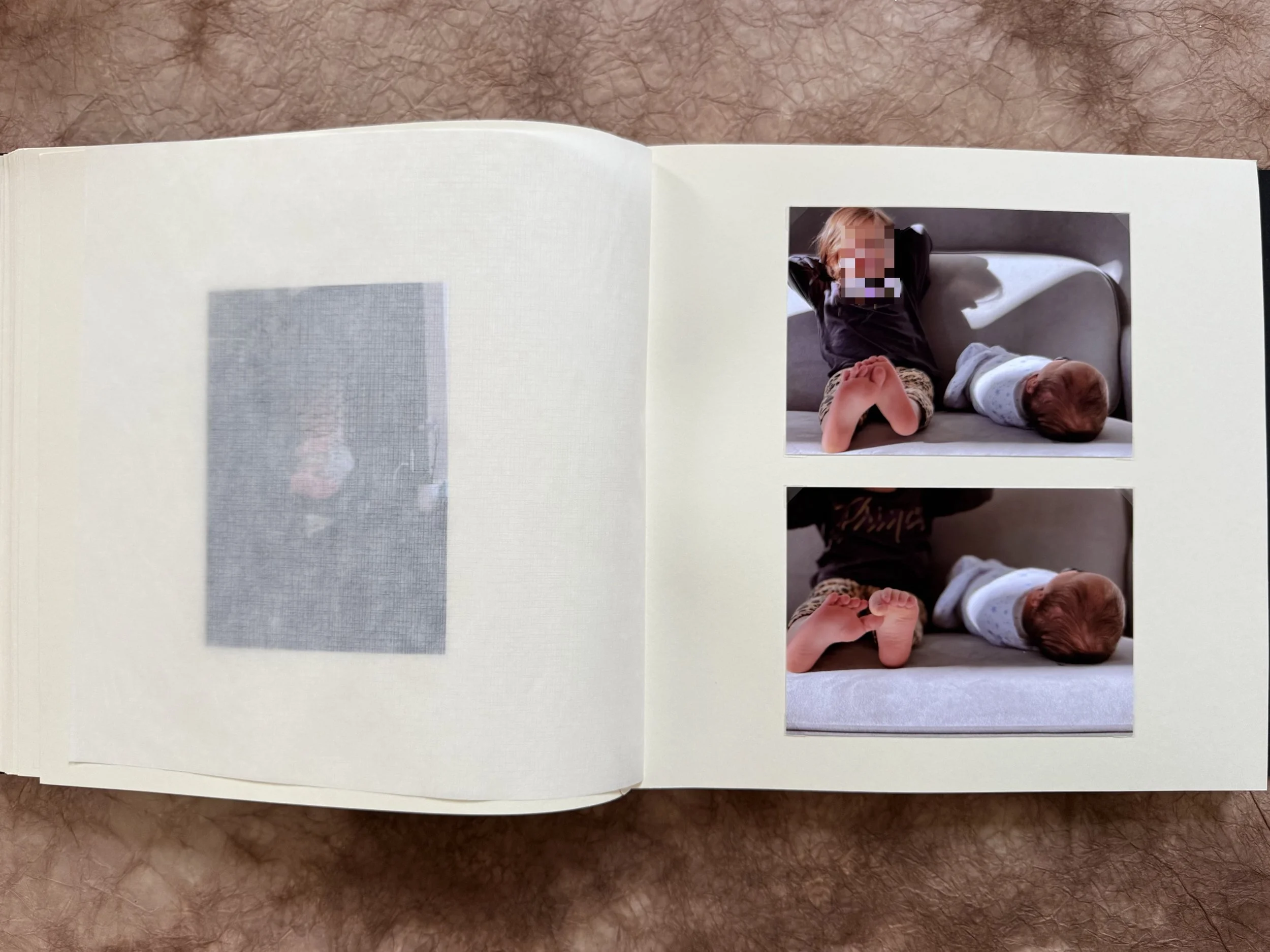 Open photo album with two photos of a sleeping baby on a couch, one of which has a pixelated face.