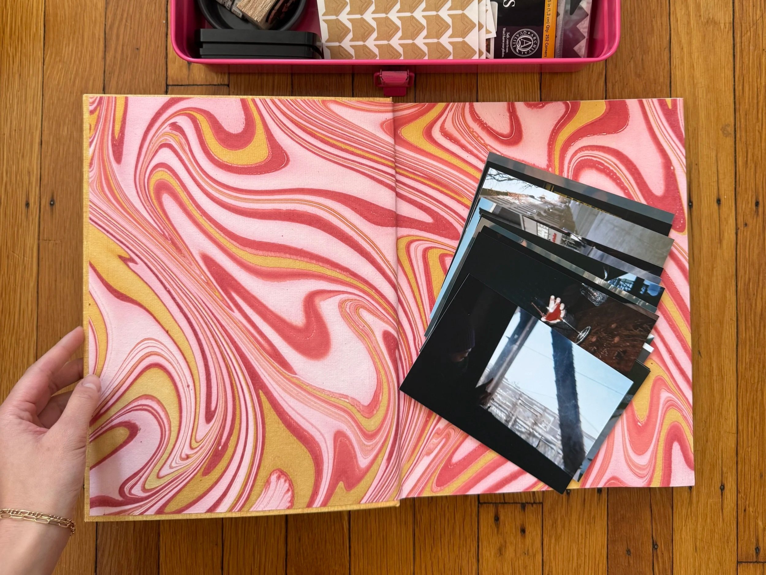 A scrapbook with a pink and yellow marbled cover, open on a wooden table, with four photographs inside. A hand is holding the scrapbook open, and there is a pink box with various supplies above it.