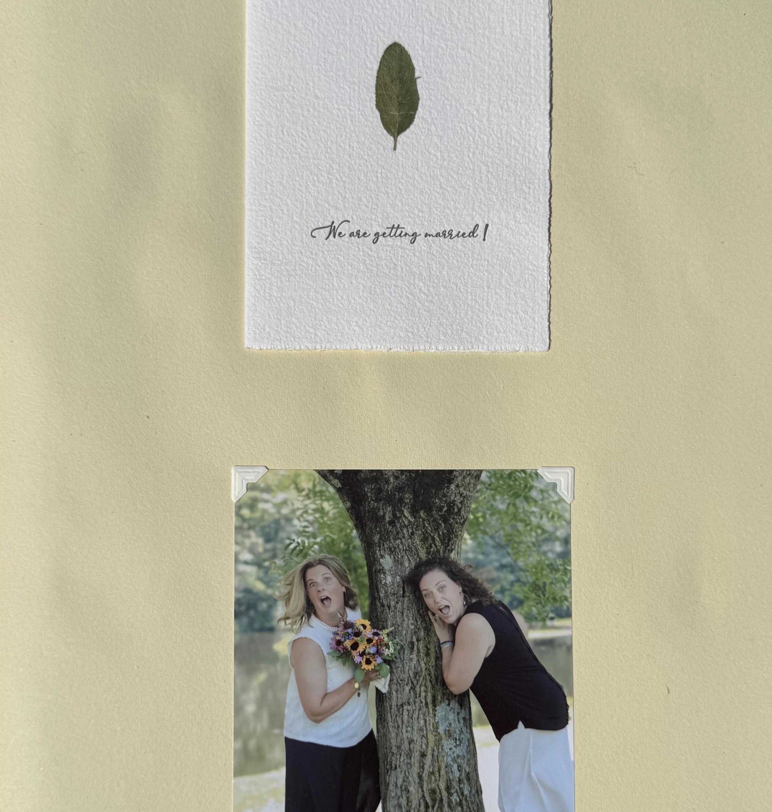 A wedding invitation with a pressed leaf and the message 'We are getting married!' above a photo of two women making playful faces at a tree by a river, one holding a bouquet of flowers.