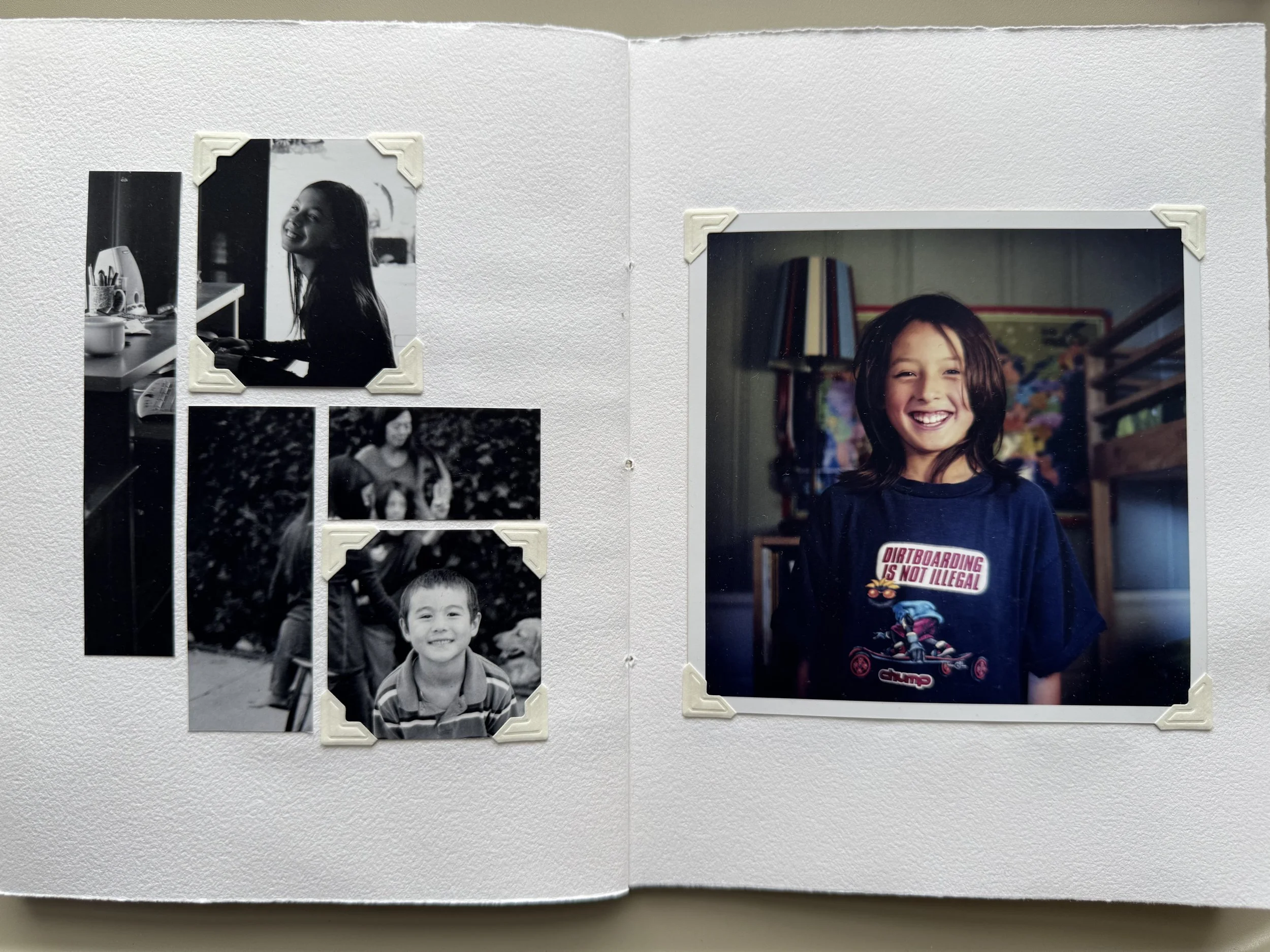 A scrapbook page with four photographs of children, three in black and white and one in color. The large color photo depicts a young girl smiling and wearing a dark T-shirt that says "Dartboarding is not illegal" with a cartoonish illustration. The b