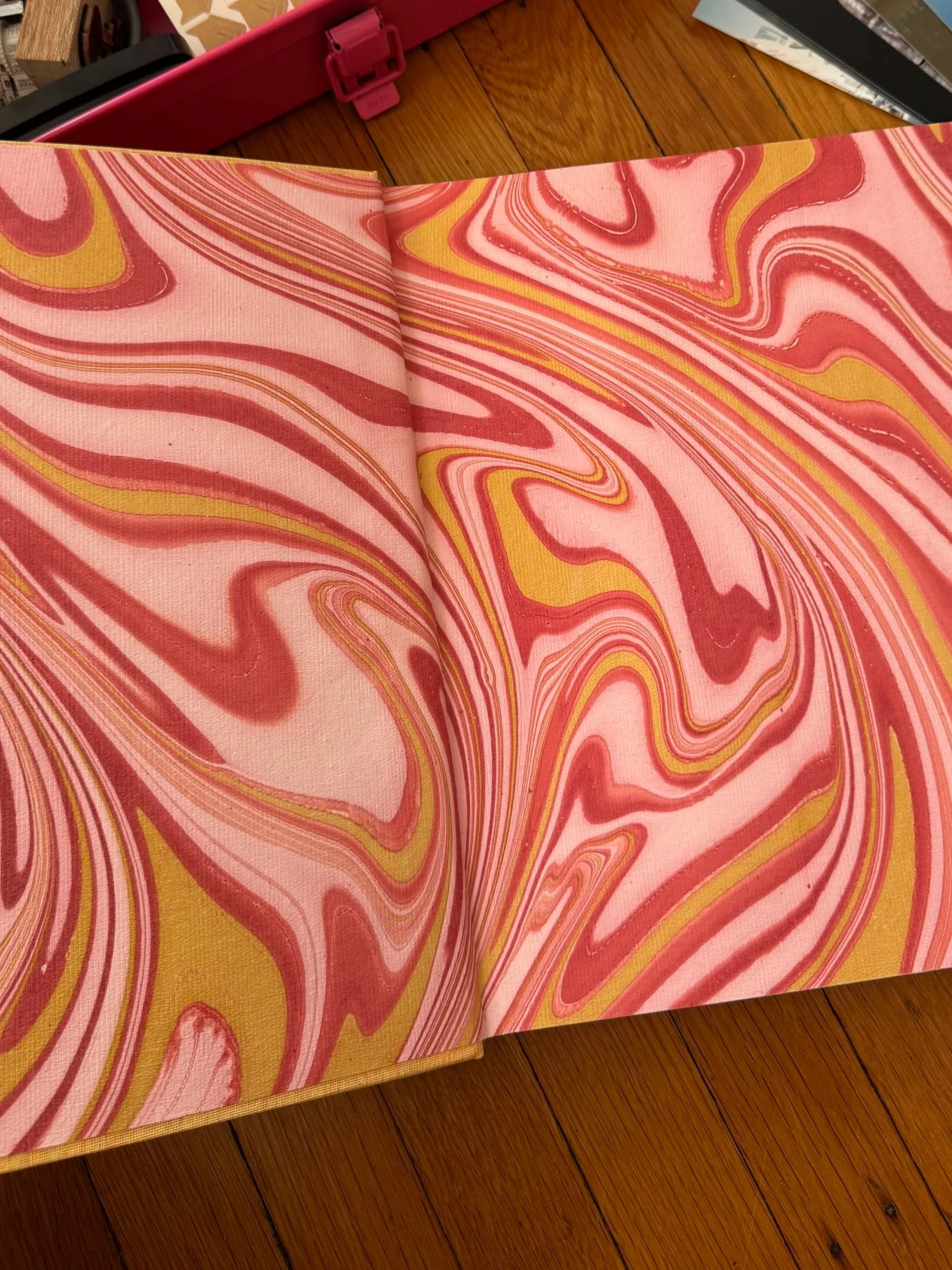 Open book revealing pink, yellow, and red marbled pages placed on a wooden floor.