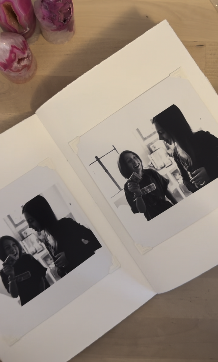 Open photo album displaying two black-and-white photos of two women, one holding a card and smiling, the other looking at her, on a wooden table with three pink candles in the background.
