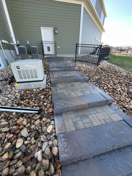 Stone pathway leading to a door with steps and a metal railing on one side, surrounded by decorative rocks with an air conditioning unit nearby.
