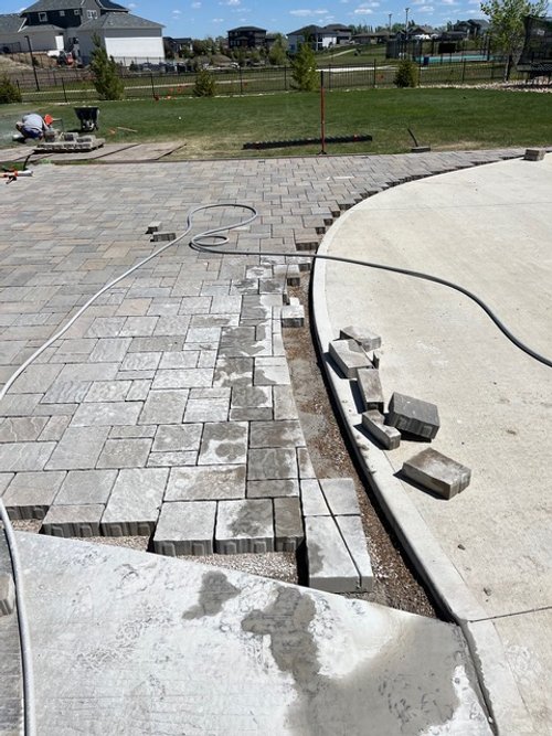 Construction site with paving stones and curbing work in progress in a backyard, with tools and materials scattered around and an orange barrier in the background.