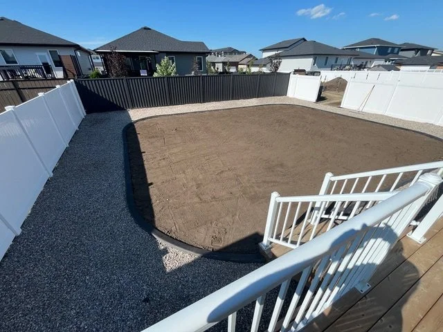 Backyard with a fenced yard, partially covered with dirt, surrounded by white and black fences, with neighboring houses in the background and a wooden deck with white stairs.