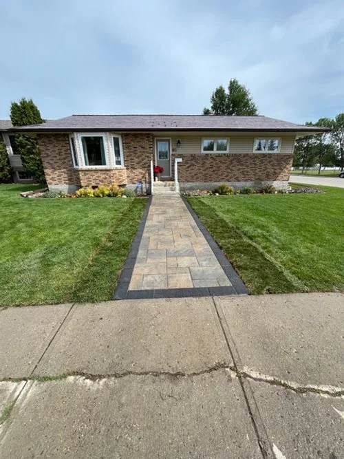 Front view of a single-story brick house with a paved walkway leading to the front door, surrounded by a green lawn, bushes, and small trees.