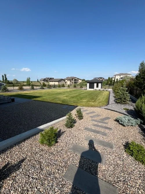 A backyard with a gravel path, small bushes, and a grassy lawn under a clear blue sky.
