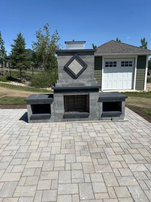 Outdoor fireplace with two side benches on a paved patio in a backyard with trees and a garage in the background.