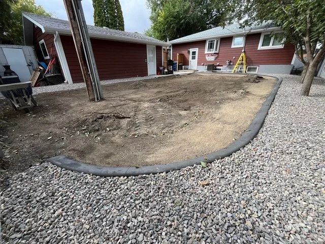 A backyard with a cleared dirt area, bordered by black edging, surrounded by gravel, with a red house, trees, and some tools and construction materials outside.