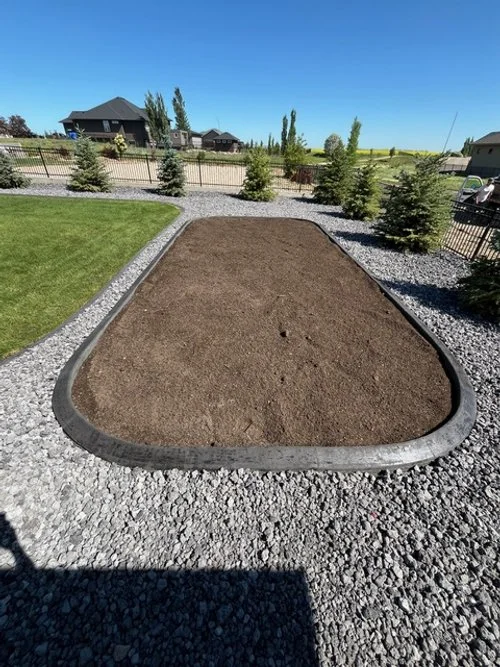 A rectangular garden bed filled with soil, bordered by a concrete edge and surrounded by gravel, with a grassy area and trees in the background.