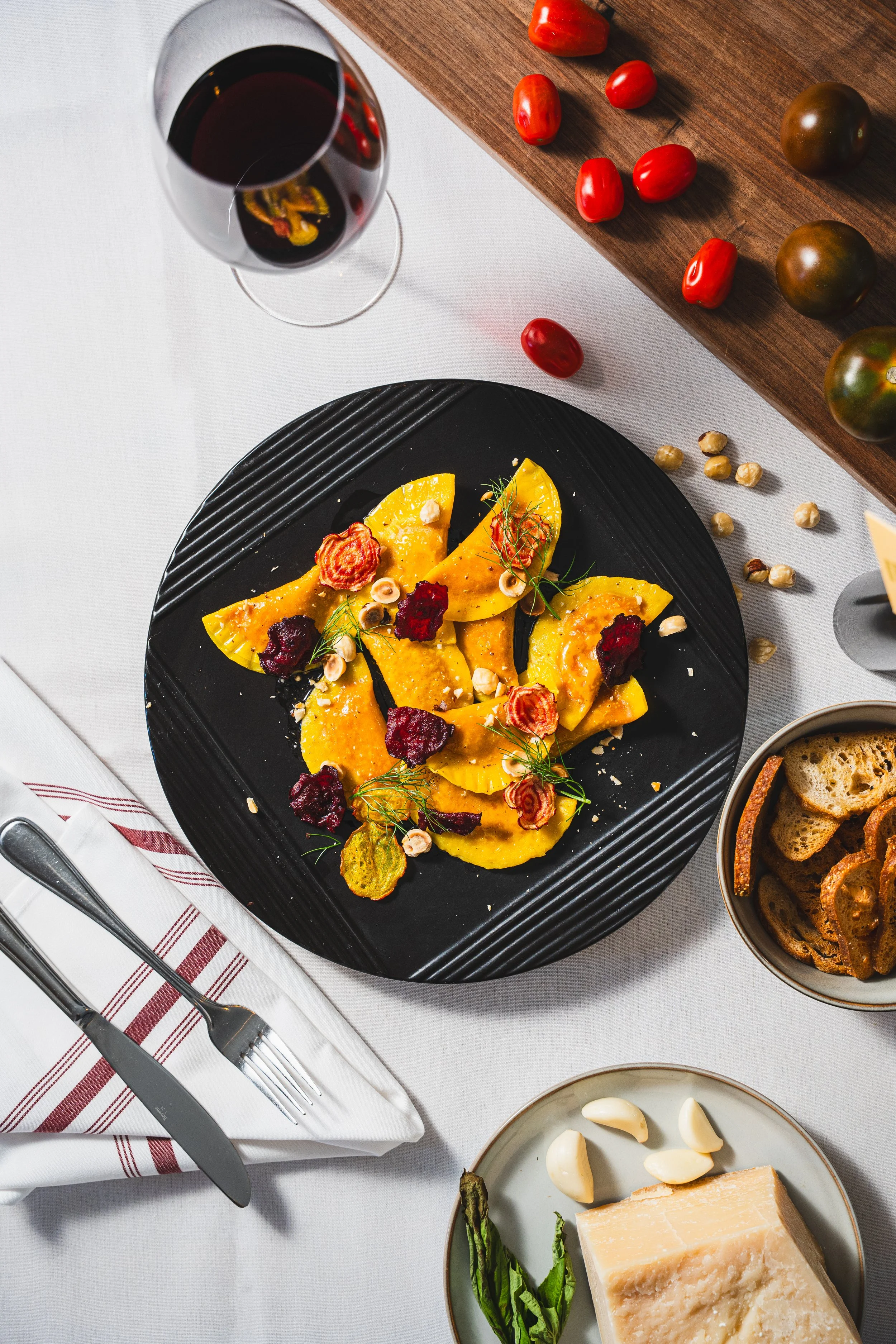 A dining table with a plate of ravioli, a glass of red wine, a small bowl of garlic, a wedge of cheese, and a cutting board with cherry tomatoes and larger tomatoes.