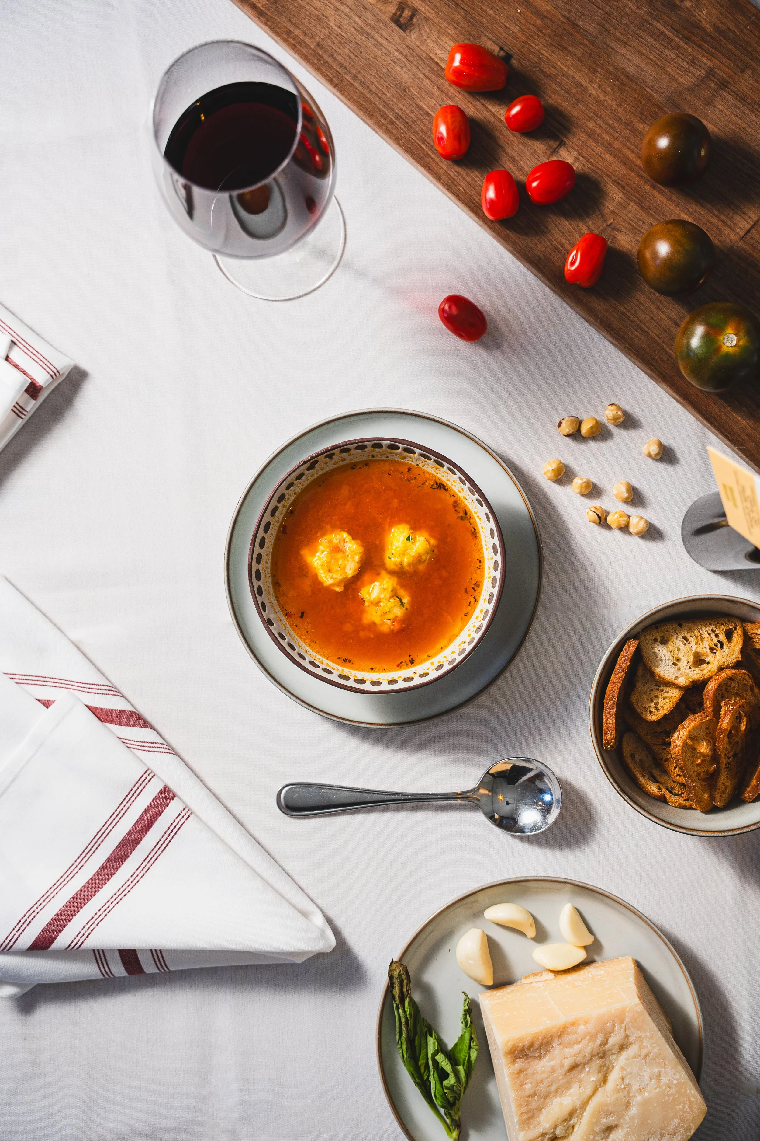 A table with a glass of red wine, a bowl of tomato soup with cheese, a bowl of crispy bread slices, garlic, cheese, basil, and a wooden board with heirloom tomatoes.