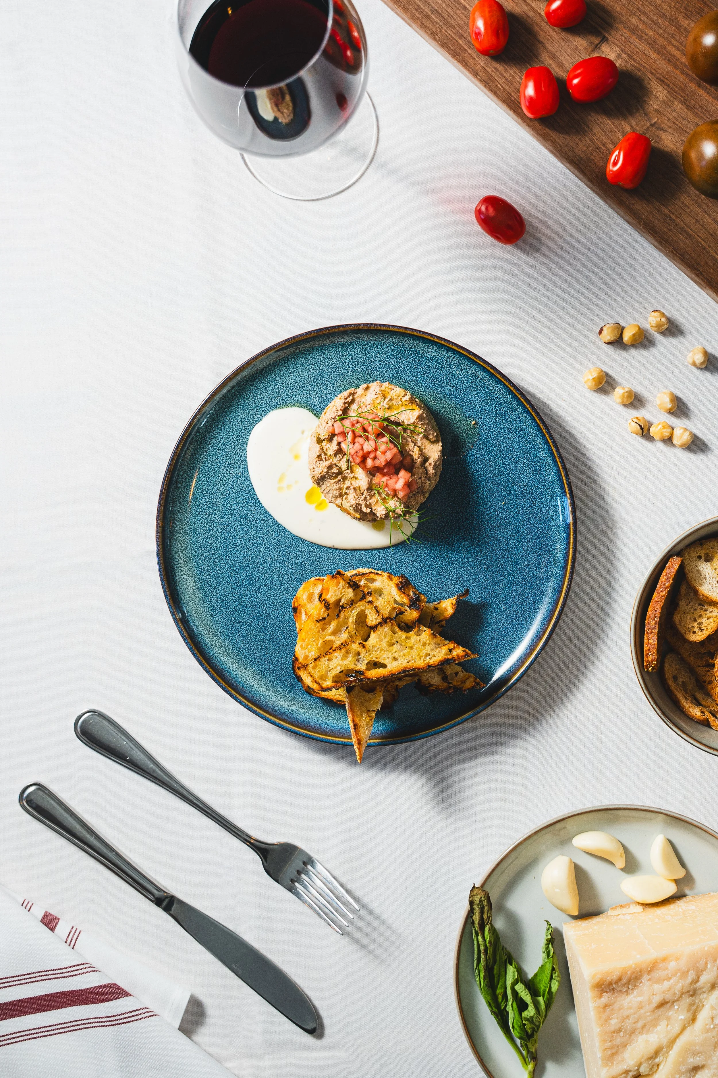 A tabletop with a blue plate containing a breaded dish garnished with diced tomatoes and herbs, and a piece of roasted bread or biscotti. There is a glass of red wine, a bowl with bread slices, a cheese piece with garlic and basil, scattered cherry tomatoes, chickpeas, and some bread slices on the side.