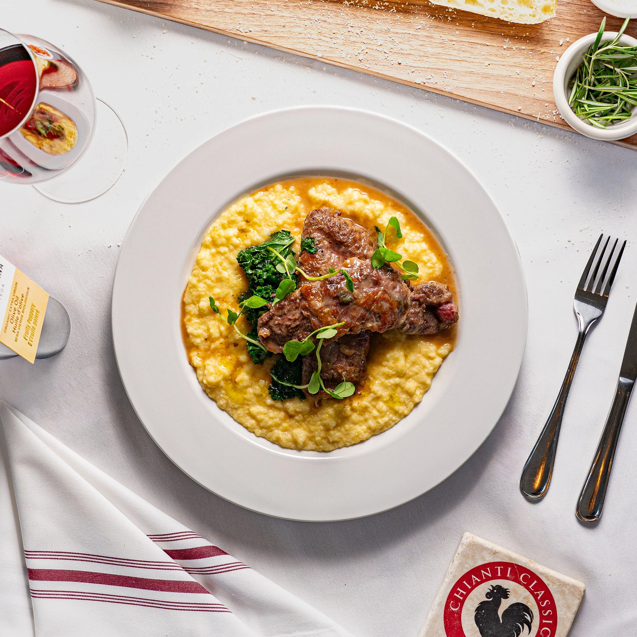 A plate of creamy polenta topped with braised beef, garnished with green herbs, with a glass of red wine on a white tablecloth.
