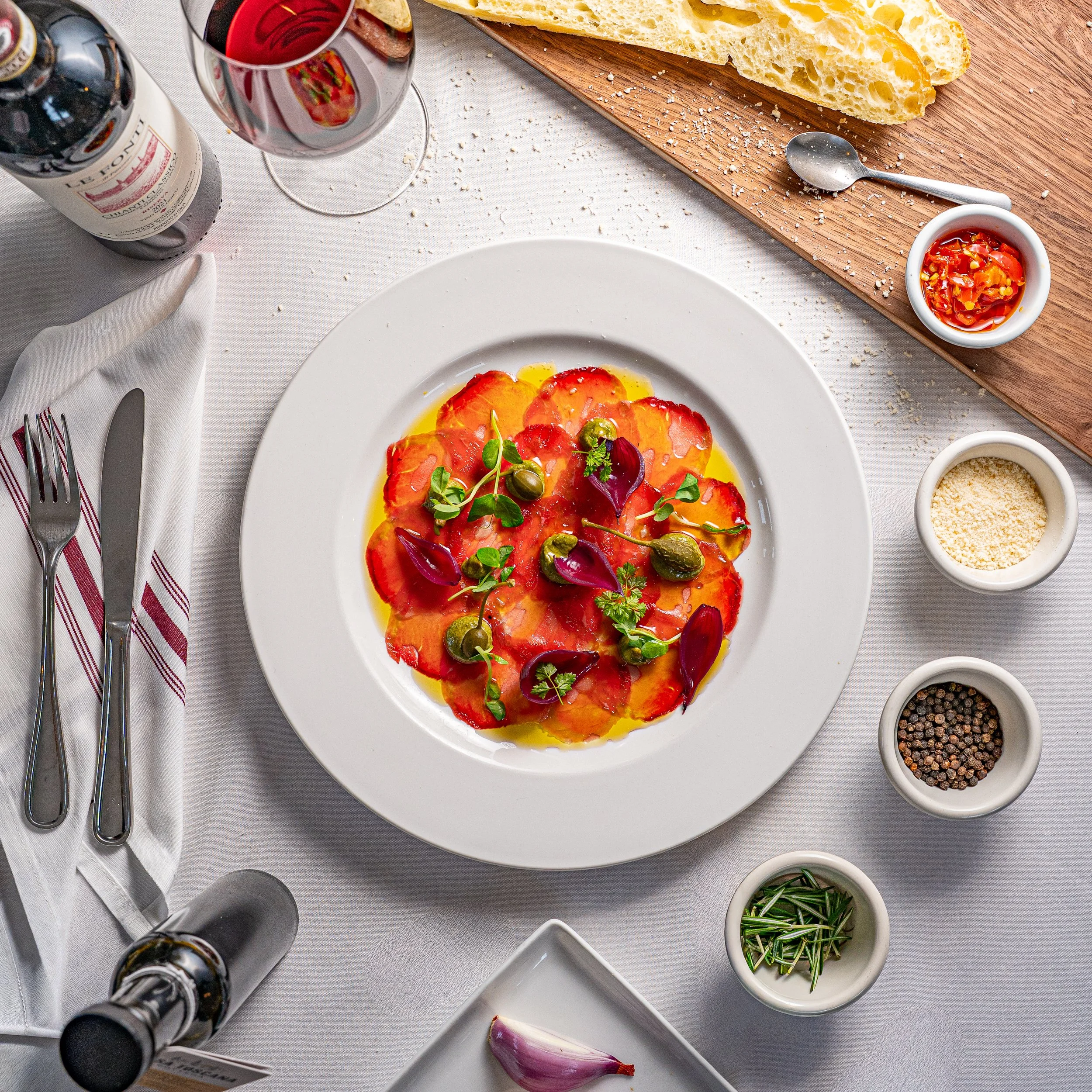 A plate of carpaccio topped with herbs, capers, and edible flowers on a white table setting with small bowls of spices, oil, and chili, a glass of red wine, a bottle, and slices of bread on a wooden board.