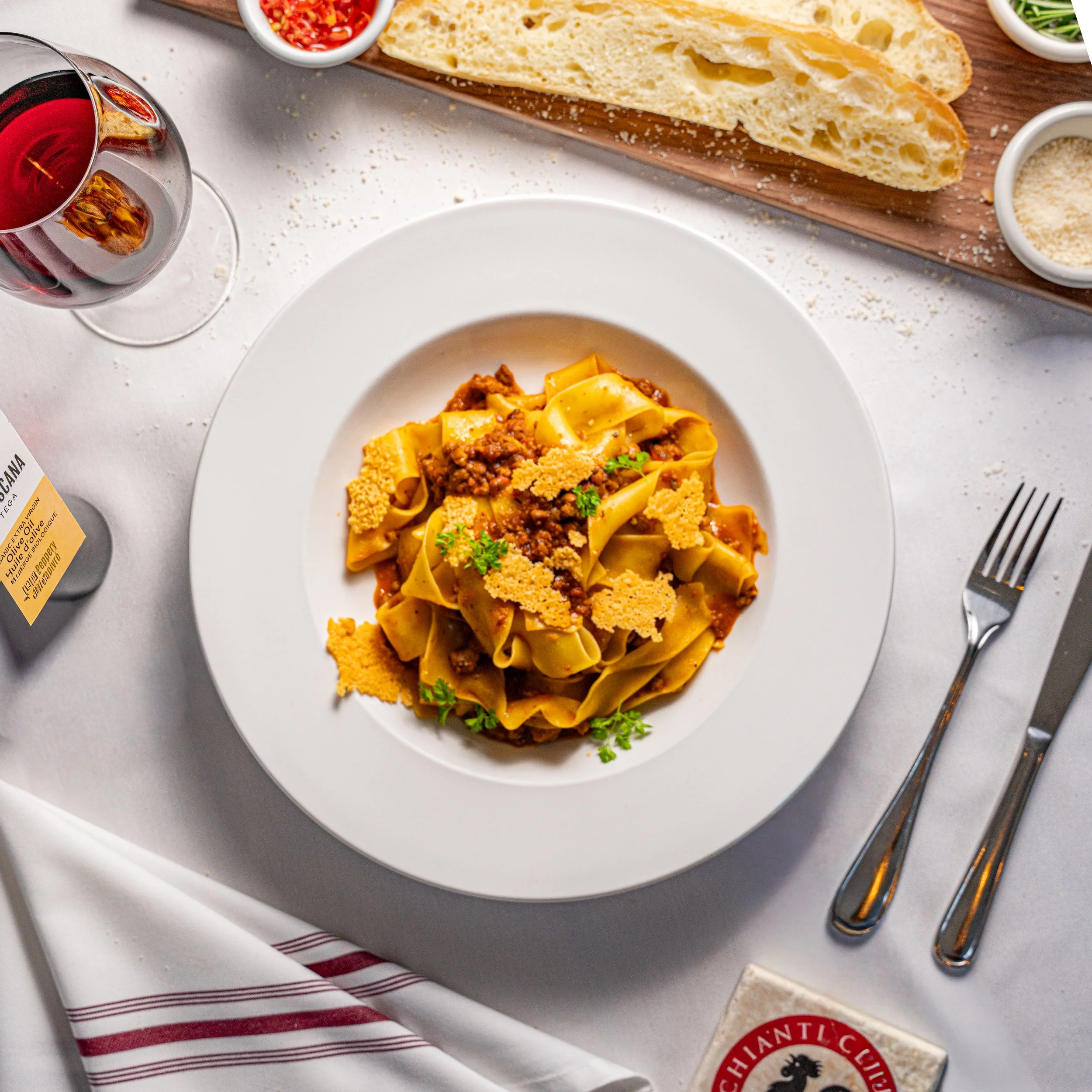 A plate of pasta with meat sauce, garnished with herbs and grated cheese, set on a white tablecloth with silverware, bread, and a glass of red wine.