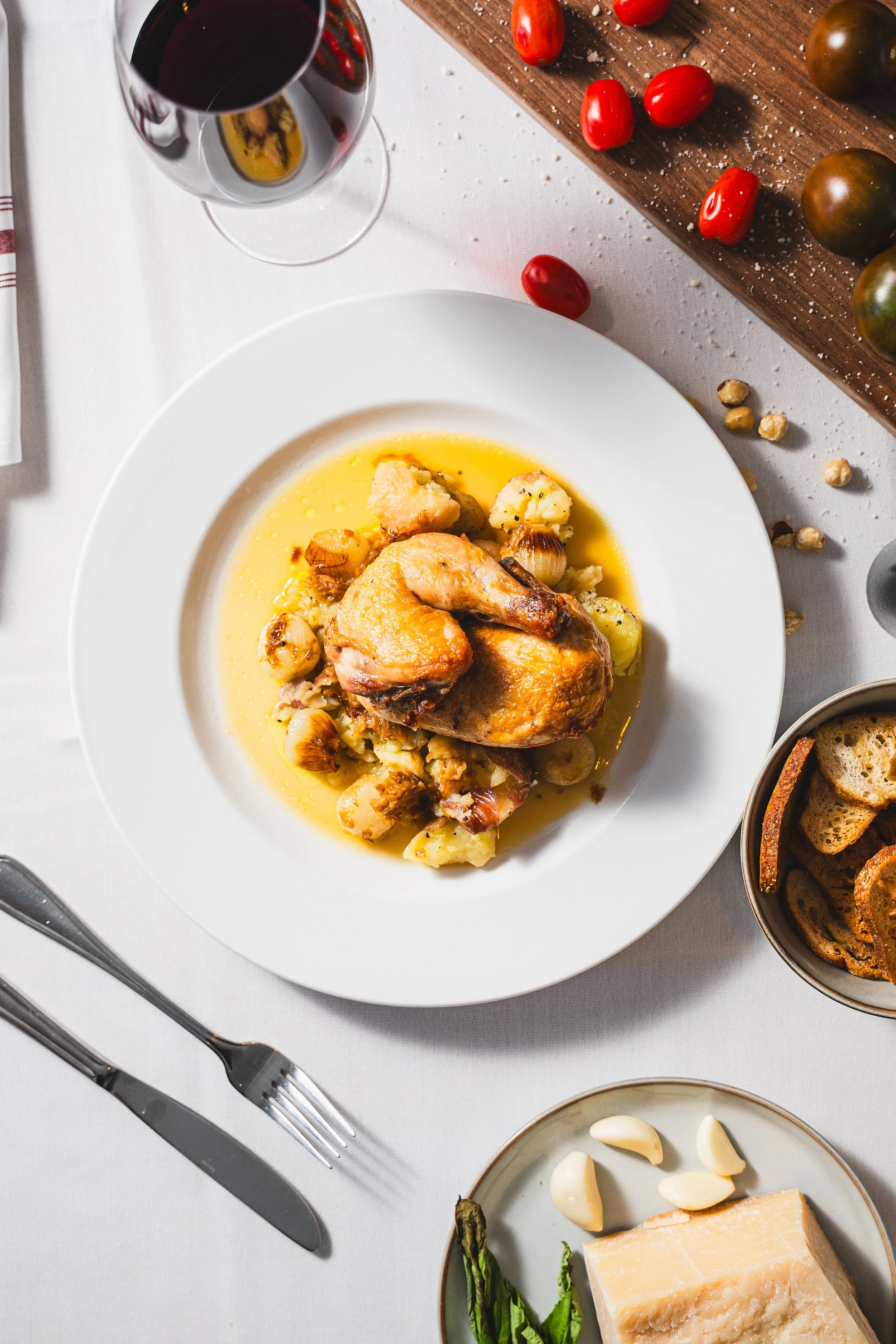 A roasted chicken leg served on a white plate with potatoes, garnished with herbs, with a glass of red wine nearby, and various vegetables and bread on the table.