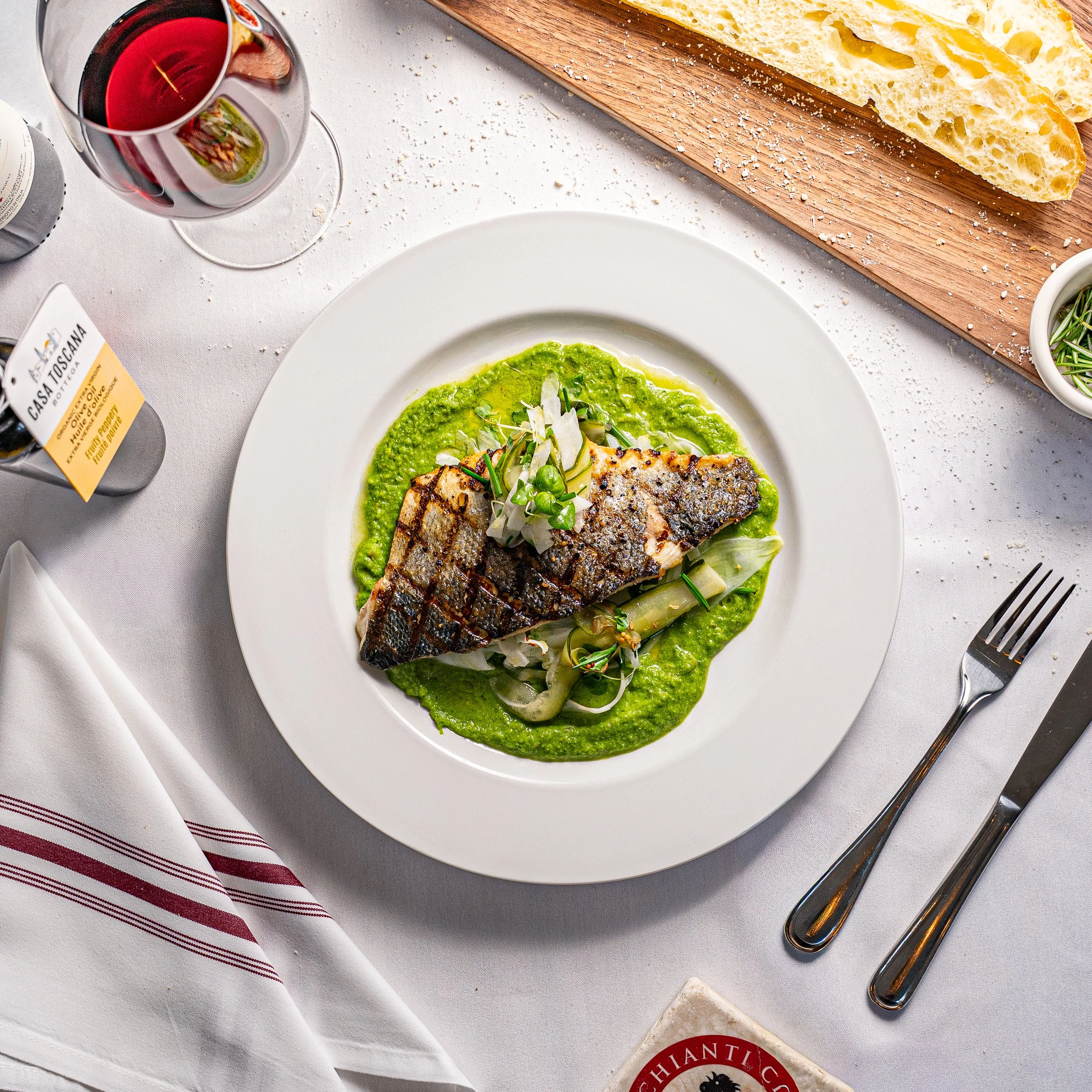 A plated dish featuring grilled fish with crispy skin, served with a green sauce and garnished with vegetables, on a white round plate. There is a glass of red wine, a wooden cutting board with bread, and some small bowls with herbs on a white tablecloth.