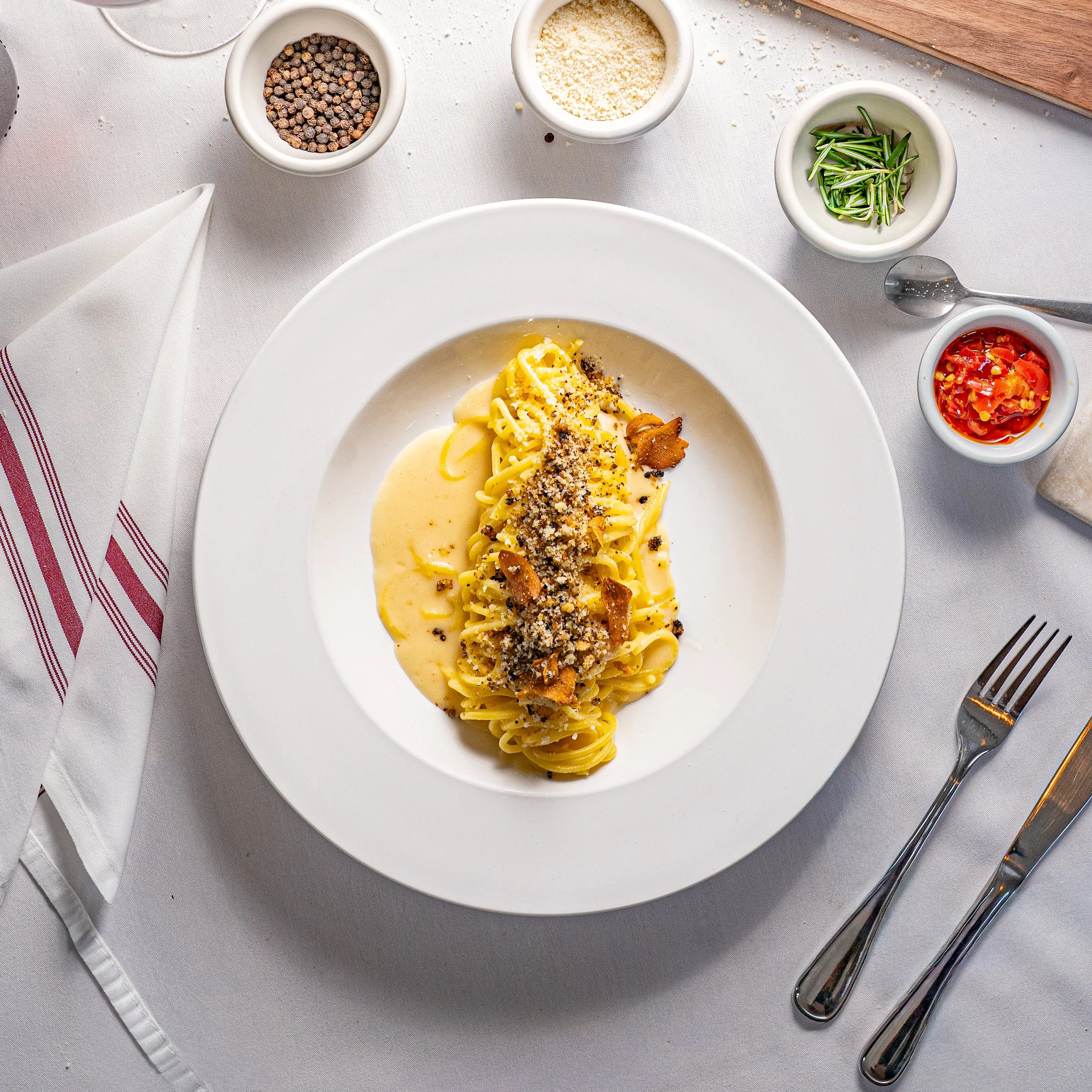 A plate of pasta with creamy sauce, topped with grated cheese and crispy bacon bits, surrounded by small bowls of pepper, grated cheese, herbs, chili peppers, and a knife and fork on a white tablecloth.