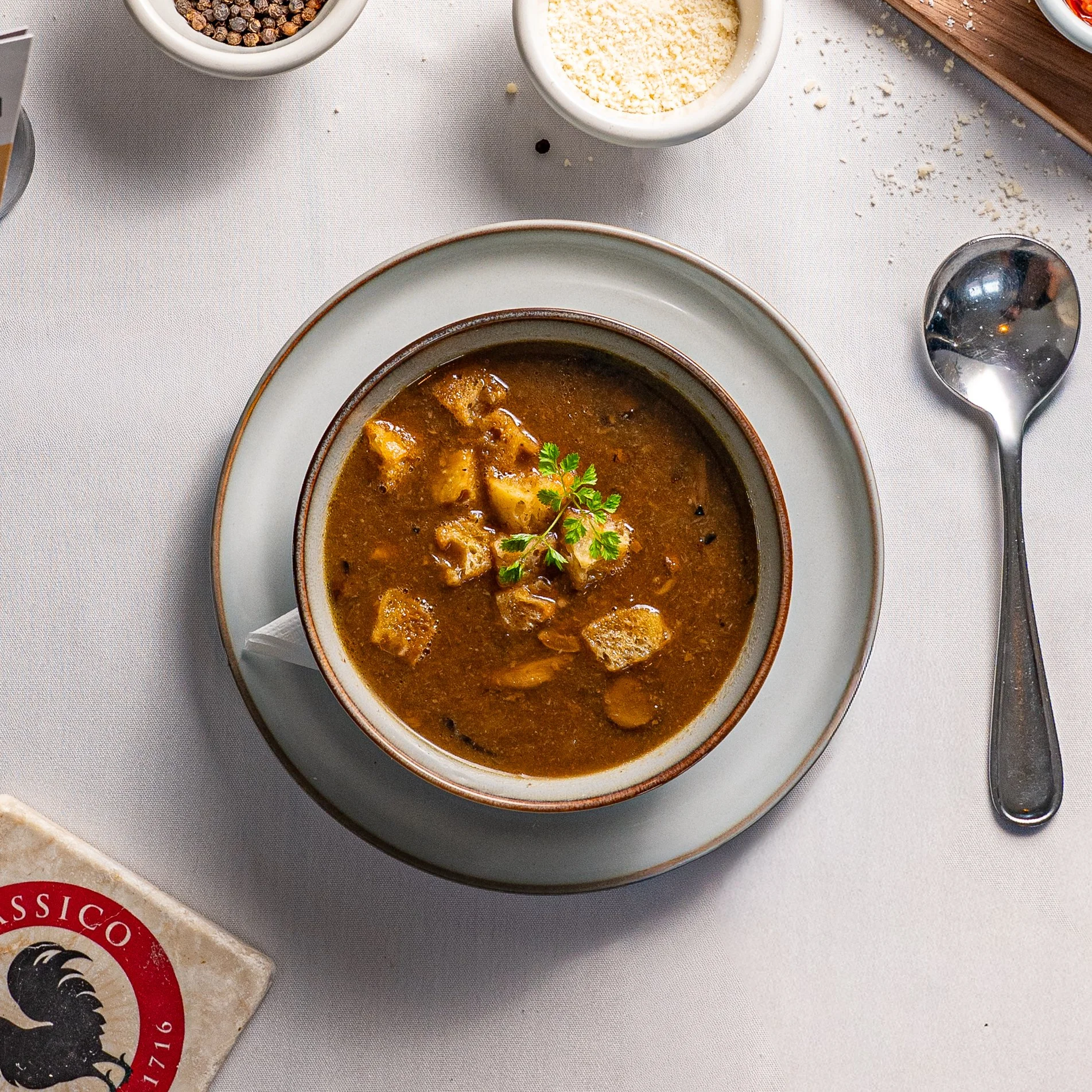 A bowl of beef stew garnished with a sprig of cilantro on a white plate, with a silver spoon beside it on a white tablecloth, along with bowls of spices and cheese around.