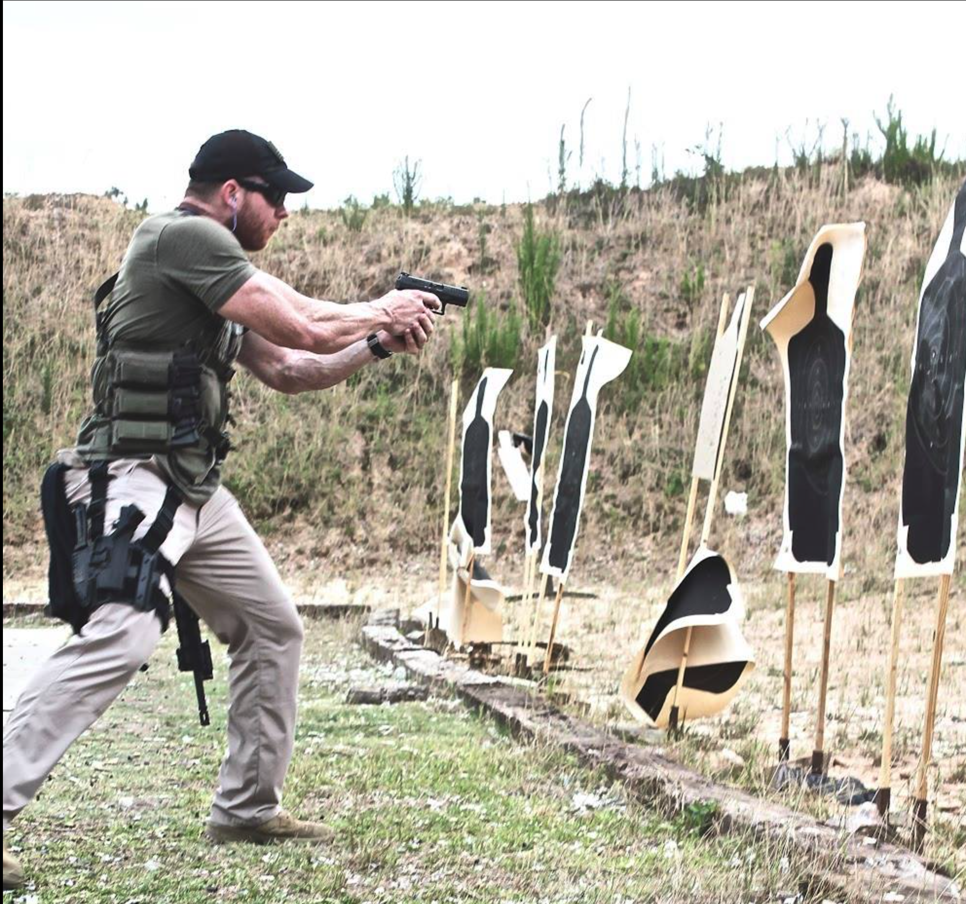 A man practicing shooting at outdoor targets with a handgun, standing in front of paper silhouettes with bullseye targets on them.