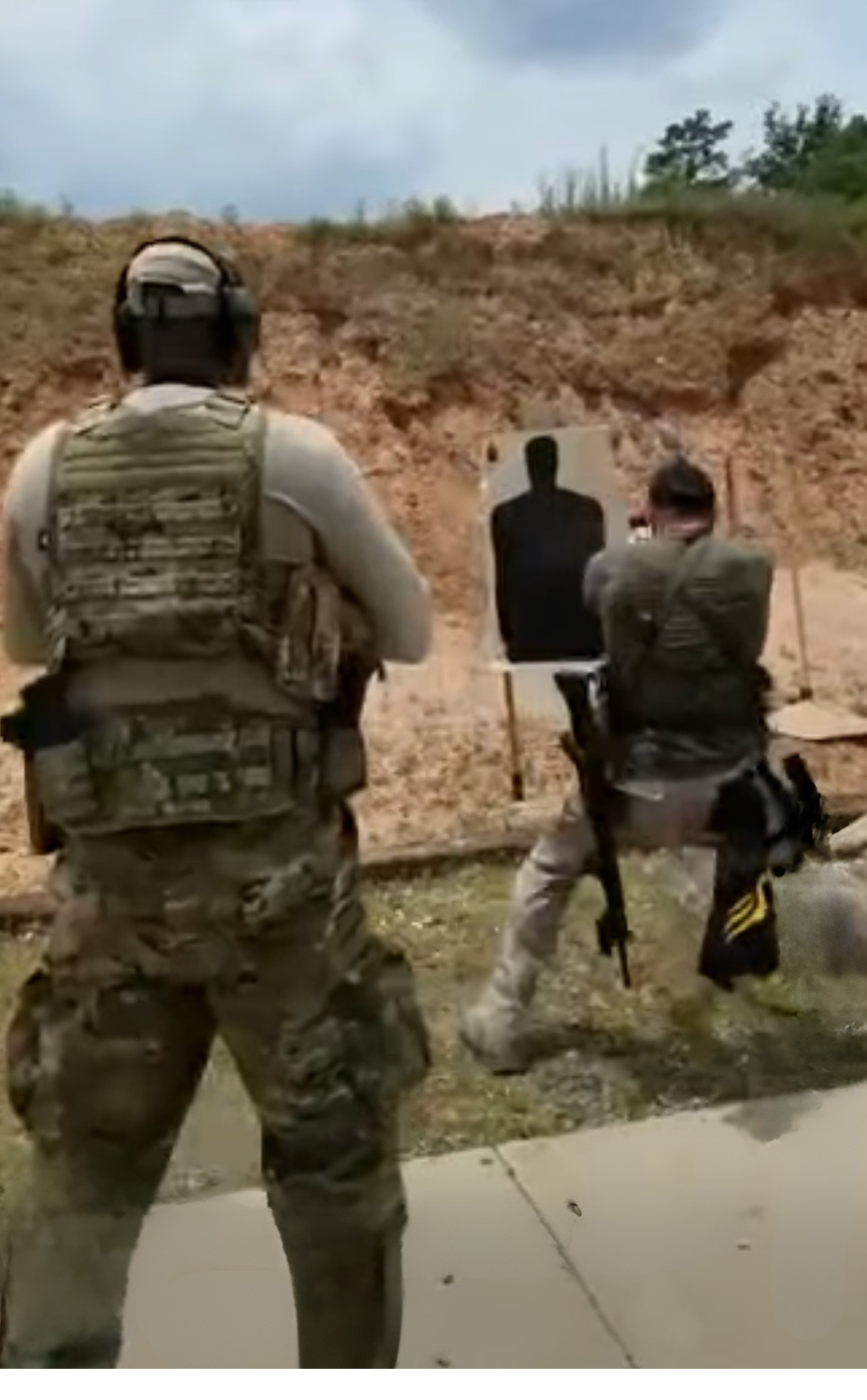 Two armed soldiers at a shooting range practicing aiming at a silhouette target, with a dirt hill and cloudy sky in the background.