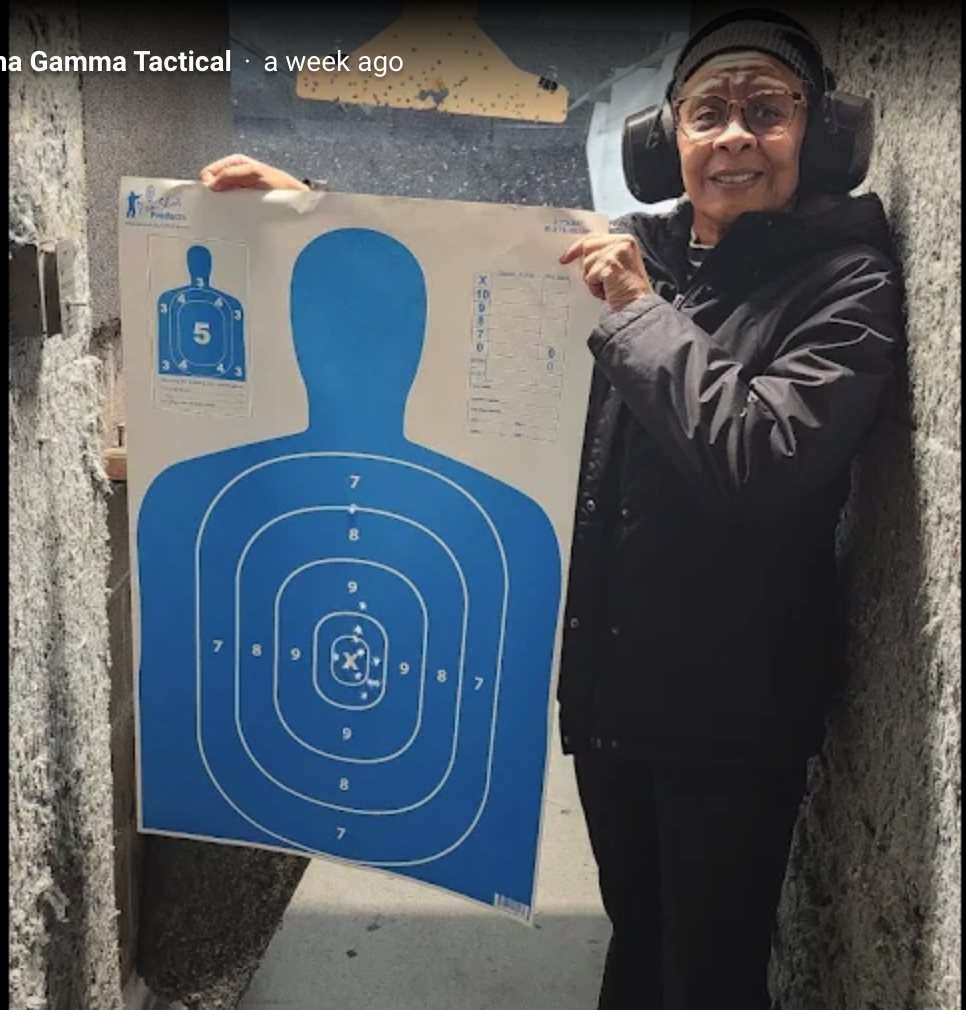An elderly woman with glasses and headphones, wearing a black jacket, smiling and pointing at a blue shooting target poster with a human silhouette, inside a shooting range or enclosed space.