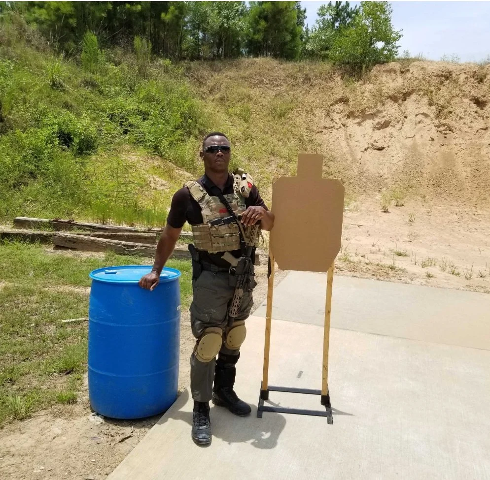 A man dressed in tactical gear standing outdoors, holding a black rifle, beside a cardboard shooting target and a blue barrel, with green trees and dirt in the background.