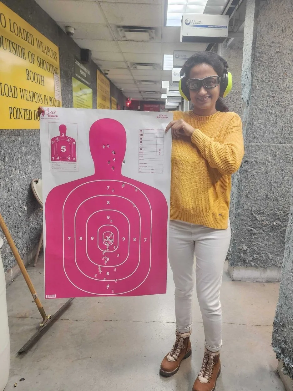 A woman at a shooting range holding a large target poster with bullet holes, smiling and wearing safety glasses and ear protection. She is dressed in a yellow sweater, white pants, and brown boots. The background shows signs with safety instructions 