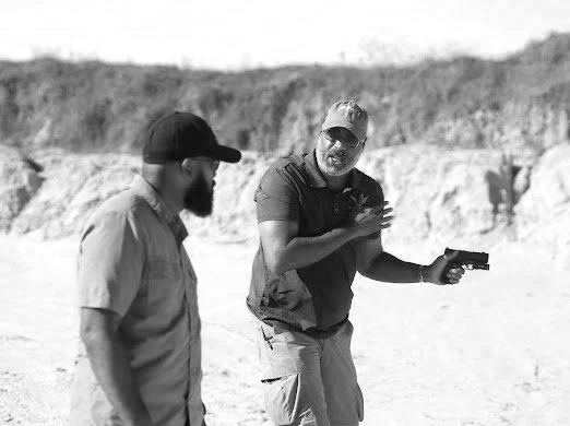 Two men in casual clothing standing outdoors near a rocky landscape, one of them holding a handgun.