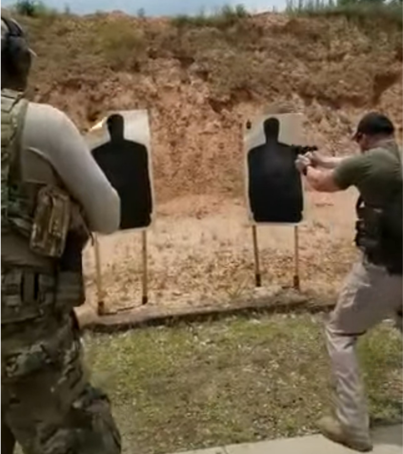 Two men practicing firearm shooting at silhouette targets positioned against a dirt wall, outdoors.