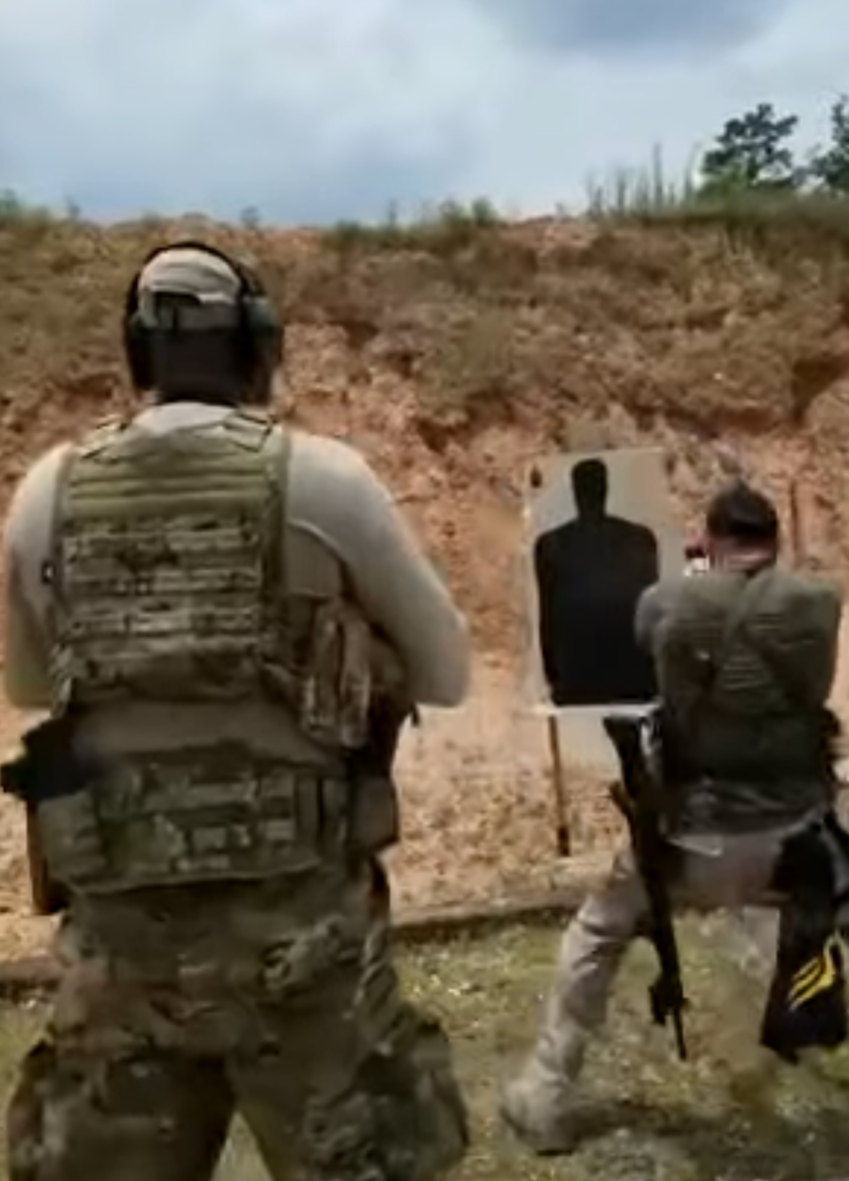 Two armed individuals in tactical gear training with a shooting target outdoors.