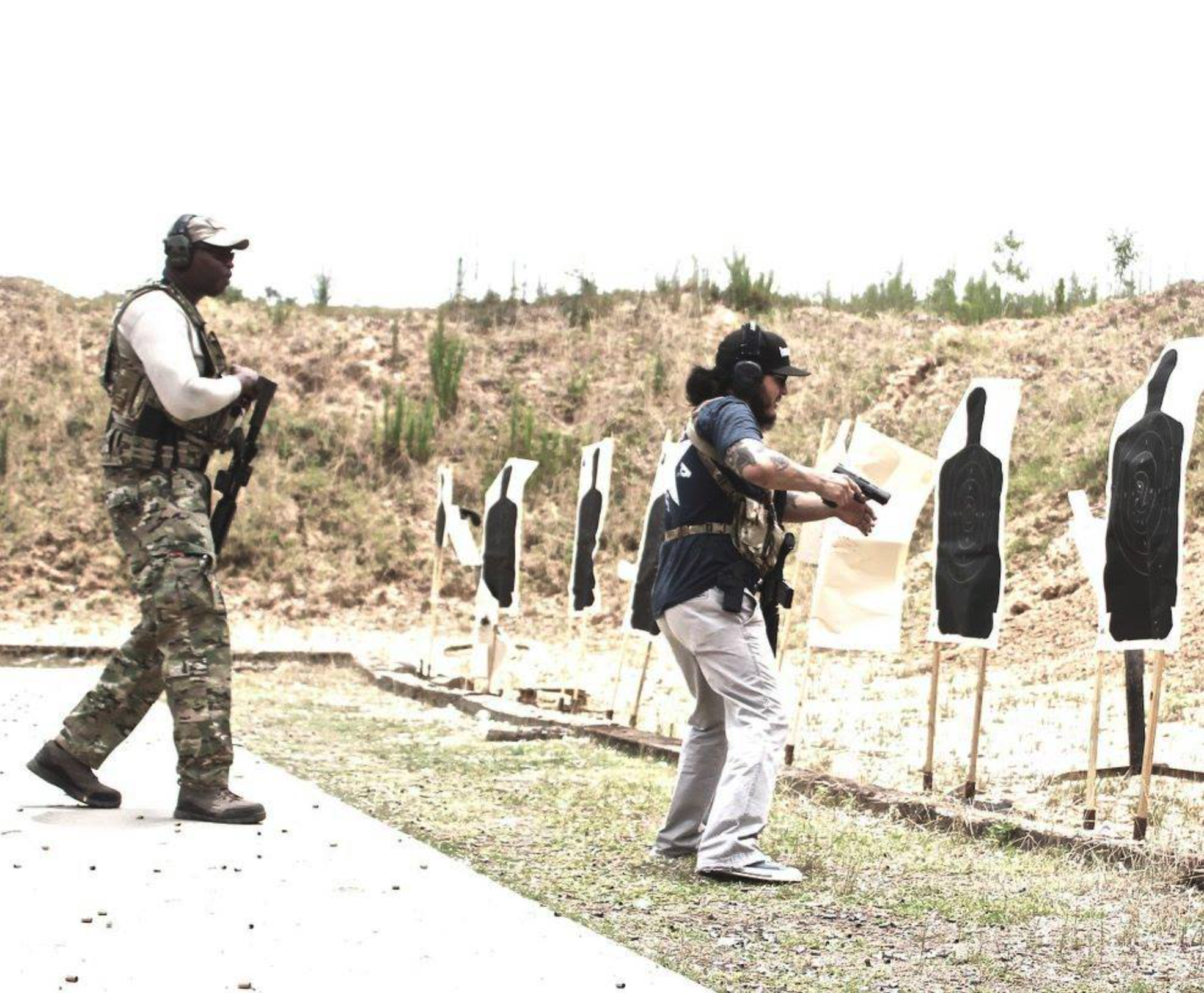 Two men practicing shooting at an outdoor shooting range with cardboard targets. One man is in camouflage with a rifle, the other man is drawing a handgun.